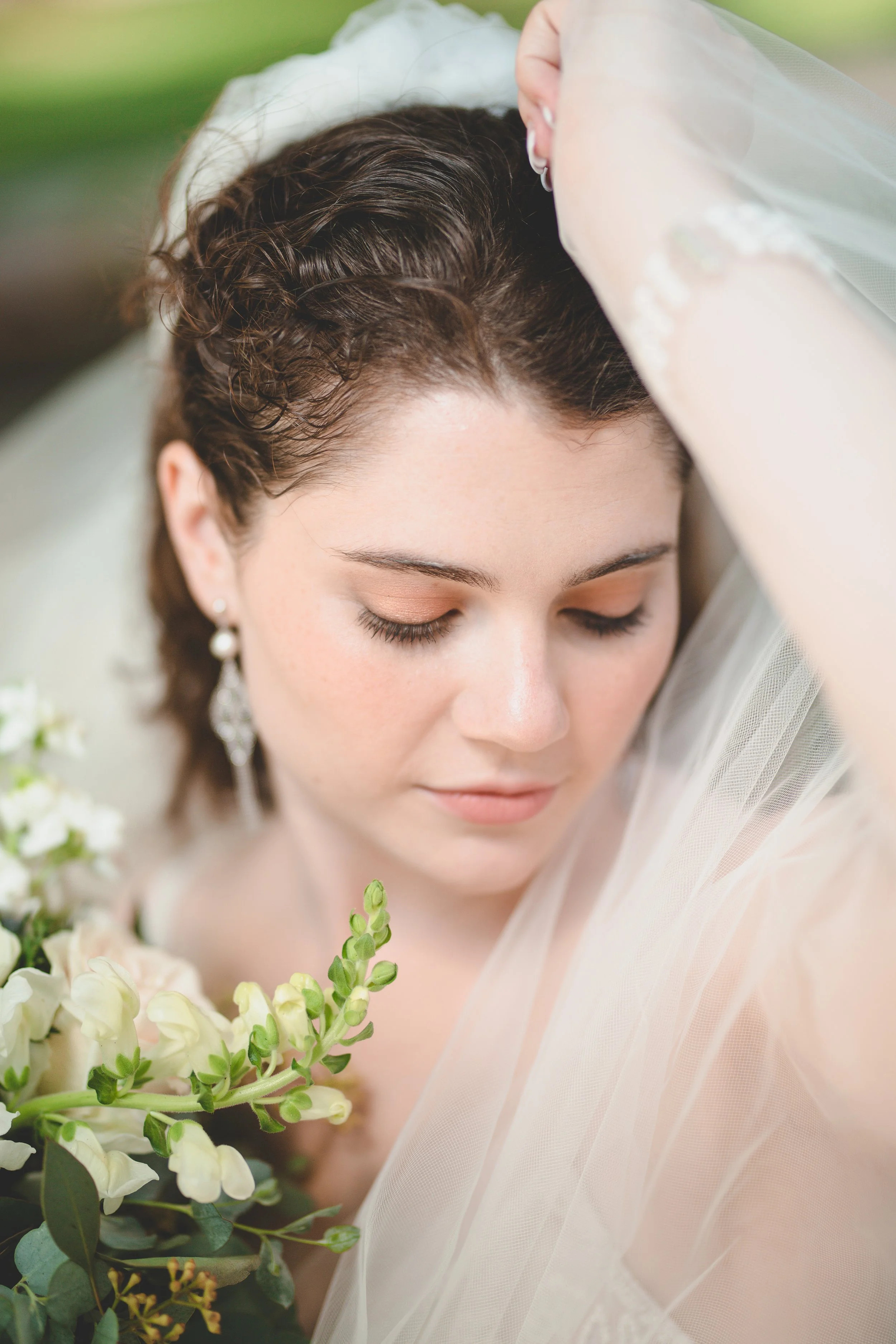 Close-up of a bride with closed eyes, holding a bouquet of white flowers, wearing earrings and a veil, with her hand resting on her forehead.