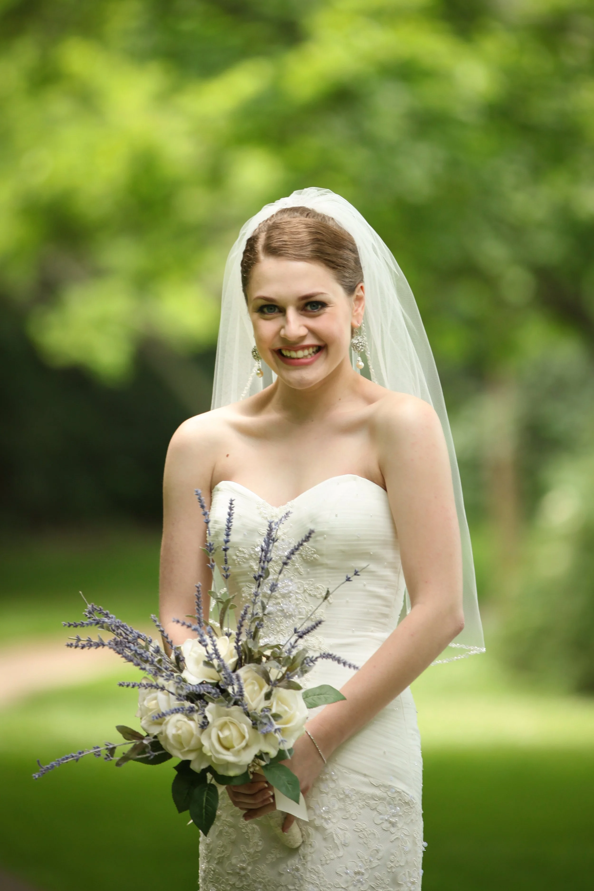 A bride in a white wedding dress holding a bouquet of white roses and purple flowers, standing outdoors with a blurred green background.