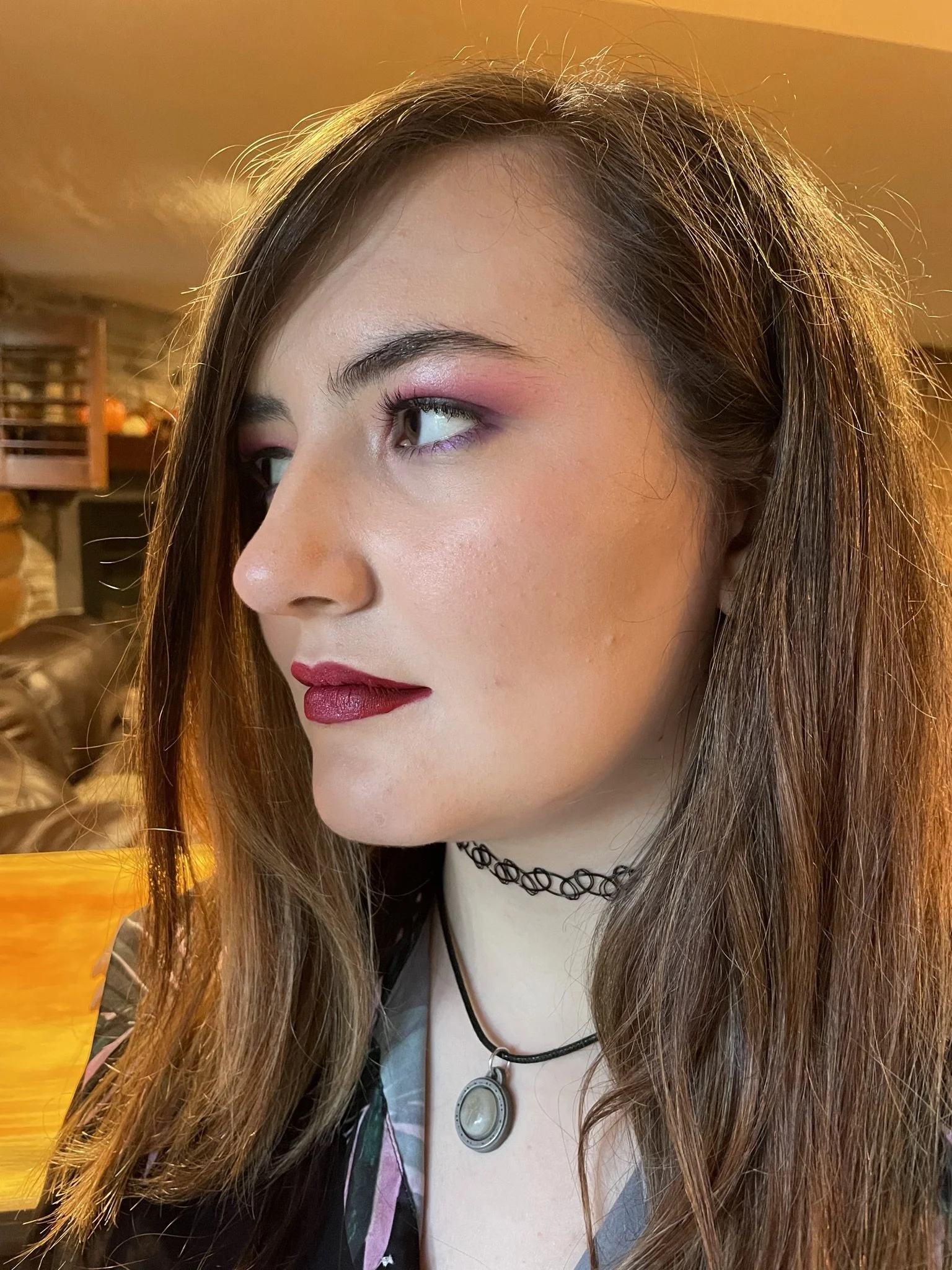 Close-up of a young woman with long brown hair, wearing pink eye makeup, dark red lipstick, a black choker, and a necklace with a pendant. She is indoors with warm lighting.