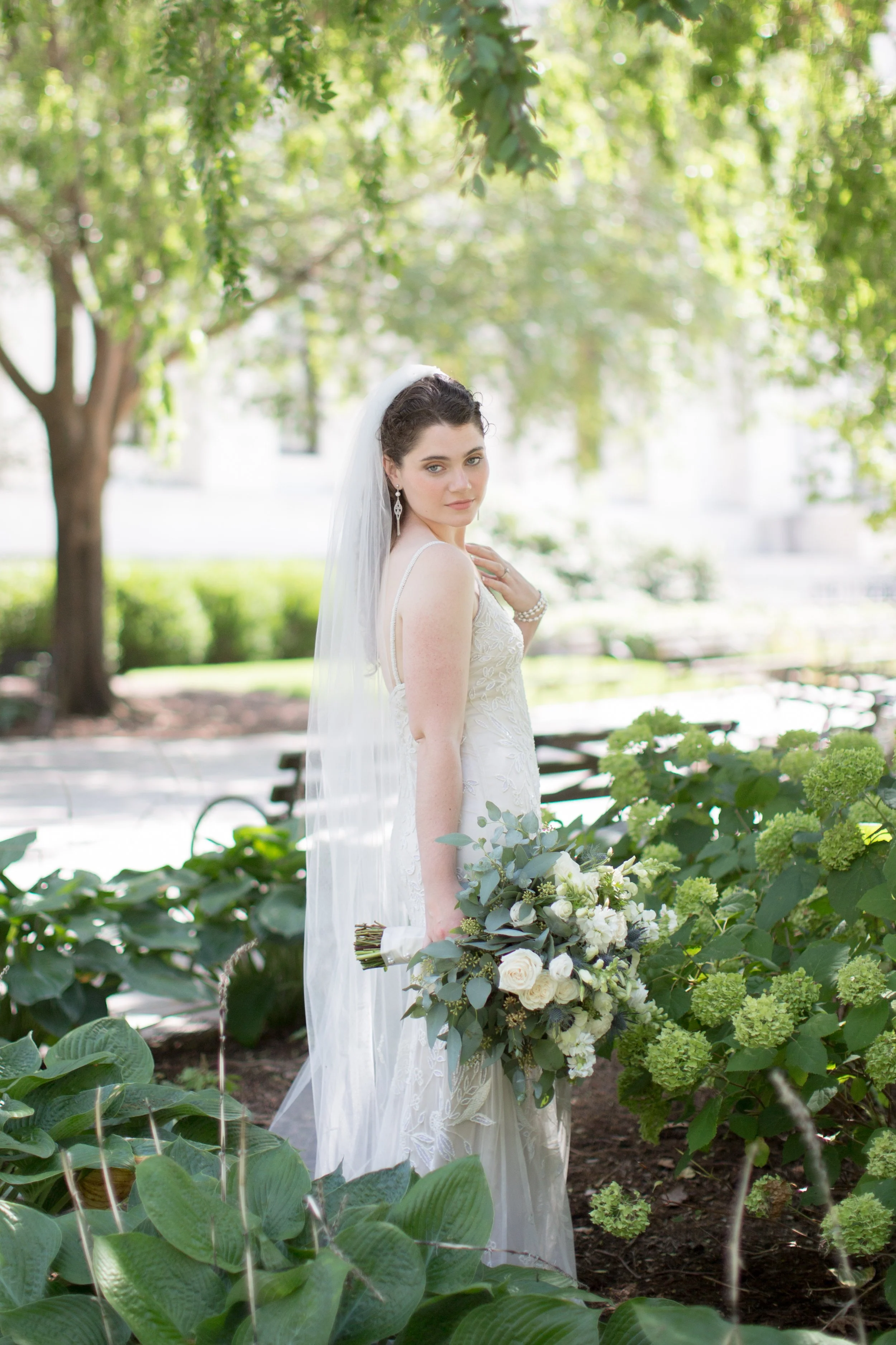 A bride standing outdoors in a park, holding a large bouquet of flowers, with trees and green foliage in the background.