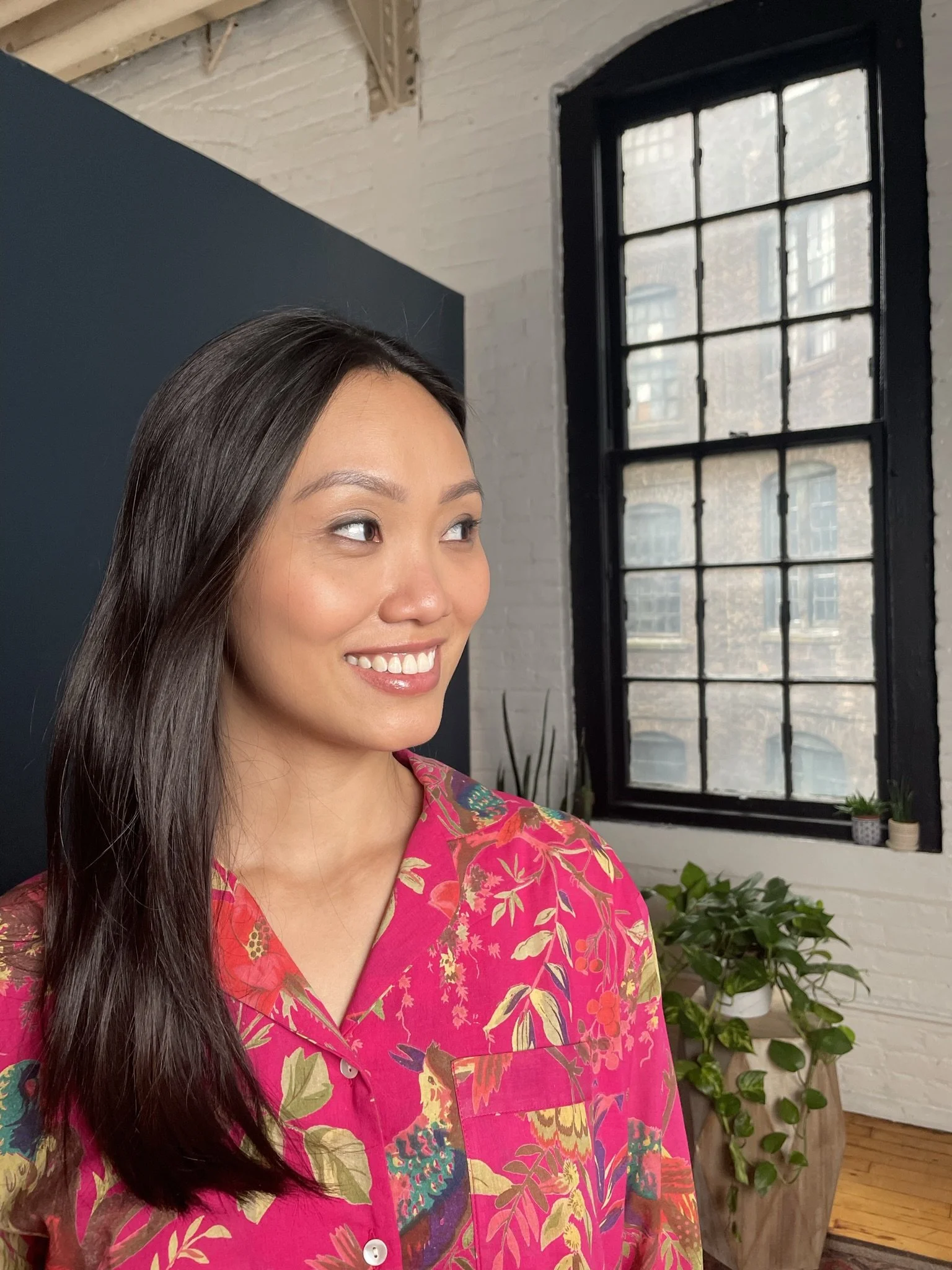 A woman with long dark hair, wearing a colorful pink floral shirt, smiling and looking to her left, in an indoor setting with a large window and potted plants in the background.