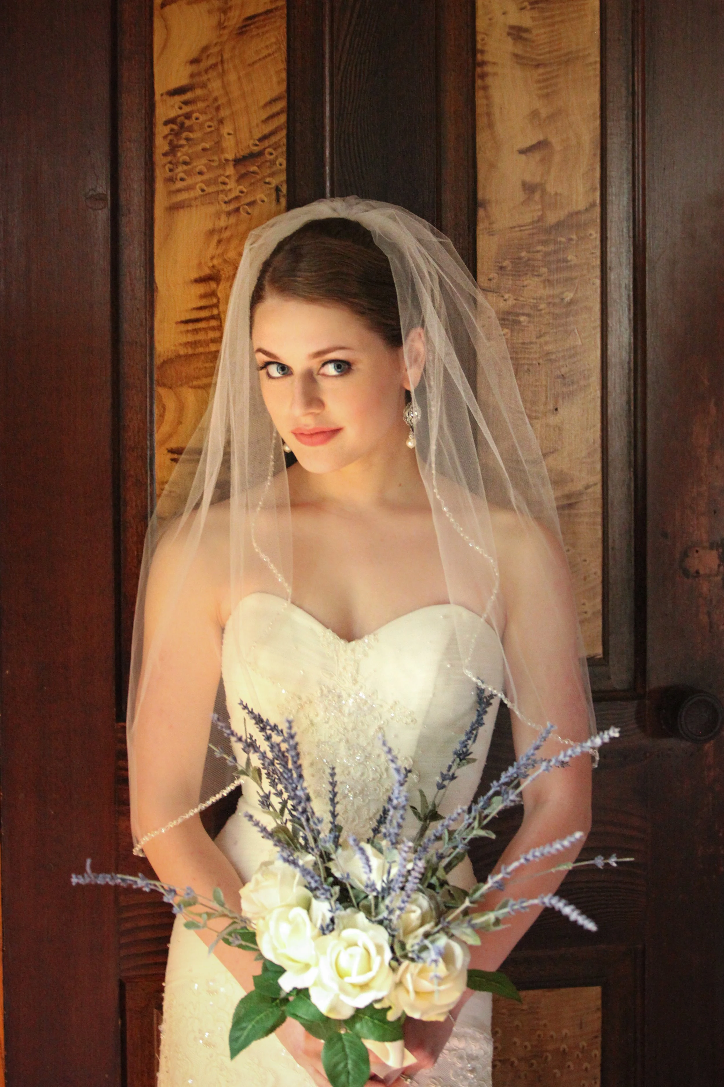 A bride with blue eyes and brown hair, wearing a white strapless wedding gown and veil, holding a bouquet of cream roses and purple lavender flowers, standing in front of a dark wooden background.
