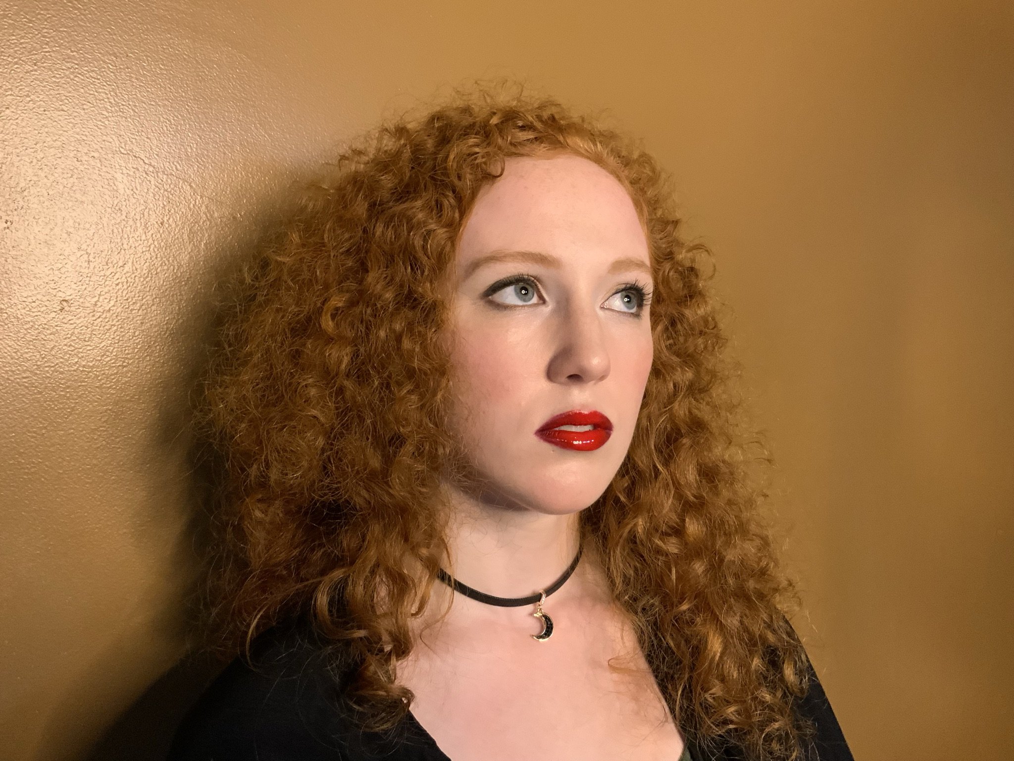 A woman with curly red hair, light skin, red lipstick, and a black choker with a moon pendant, standing against a beige wall.