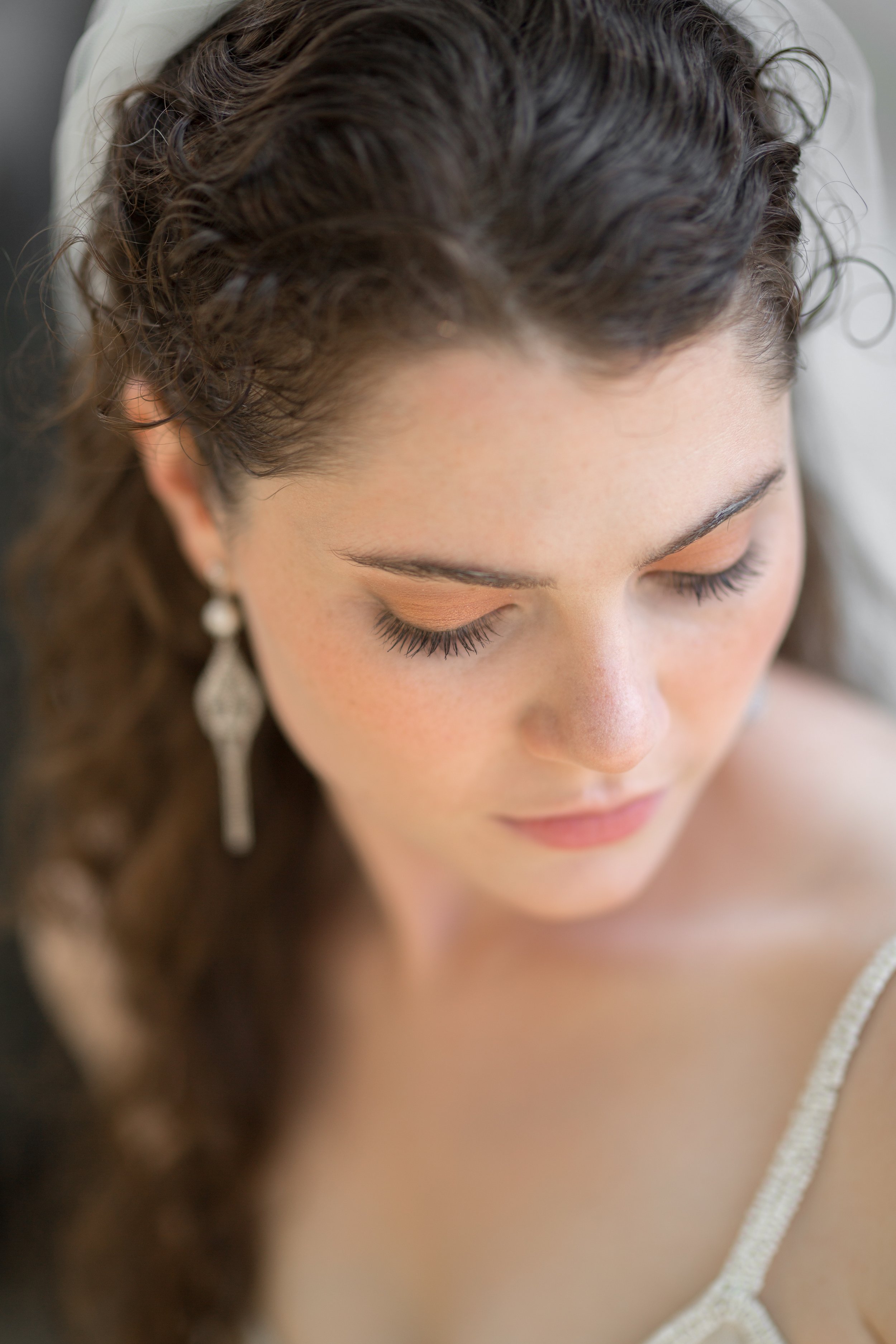 Close-up of a woman with long, dark, curly hair, wearing a white dress and dangling earrings, with her eyes closed.