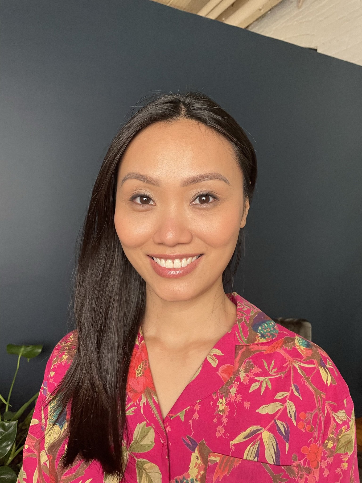 A smiling woman with long dark hair wearing a colorful pink floral shirt, standing in front of a dark blue wall with some green plants visible in the background.