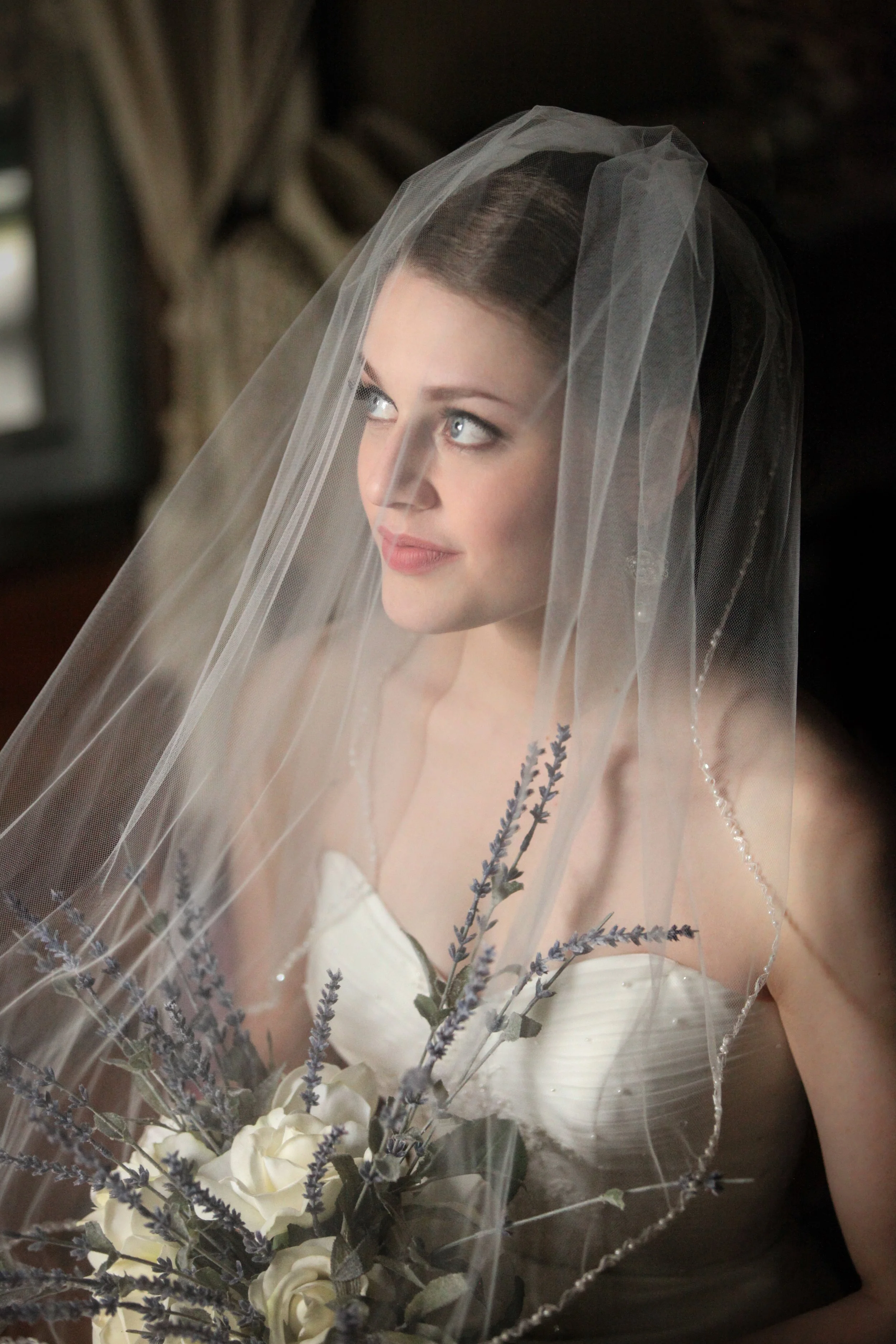 A woman in a wedding dress and veil holding a bouquet with white roses and purple flowers, sitting indoors.