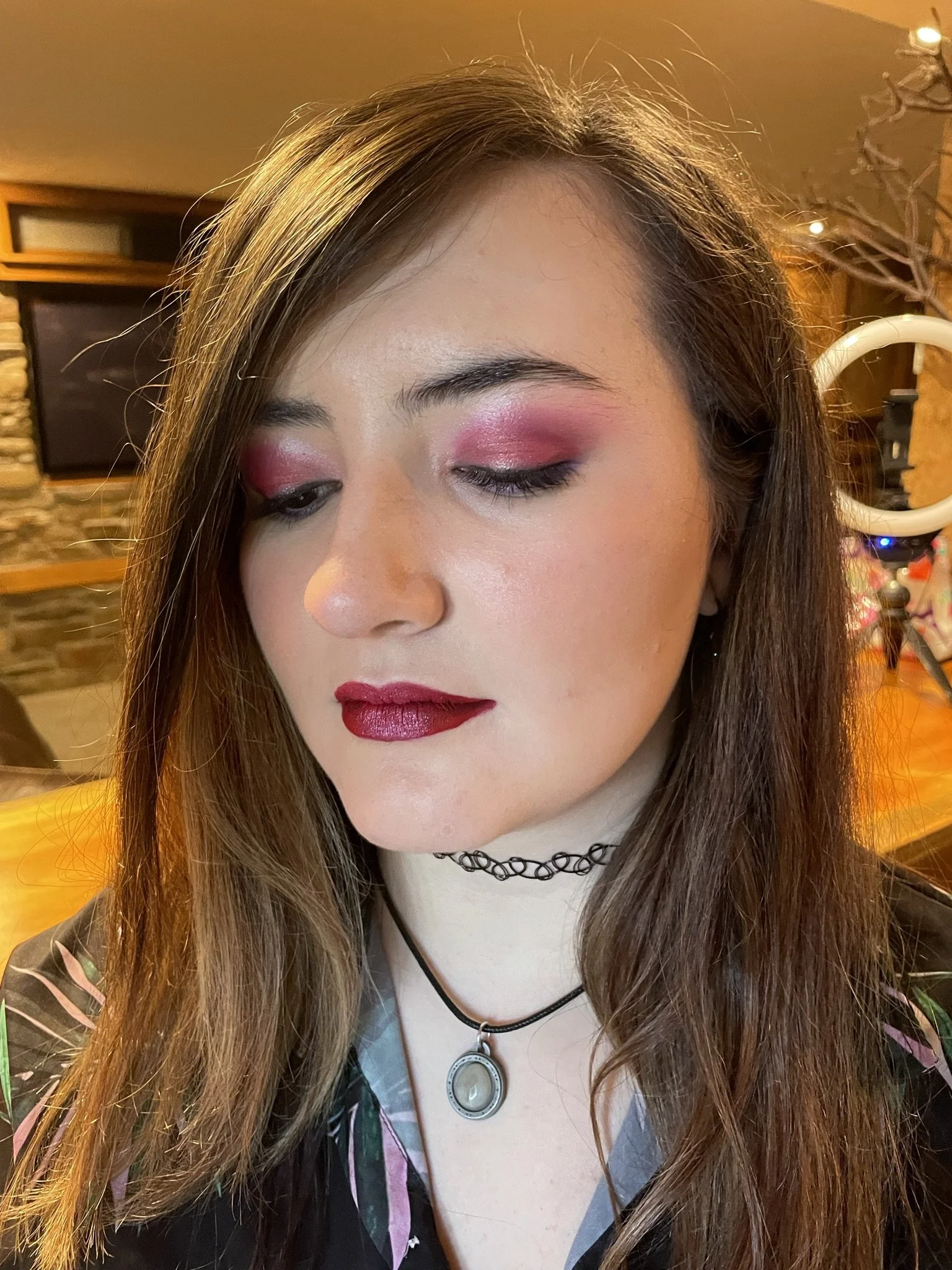A young woman with long brown hair and light skin, wearing pink eyeshadow, dark red lipstick, a black choker, and a pendant necklace, looking down with closed eyes in a cozy indoor setting.
