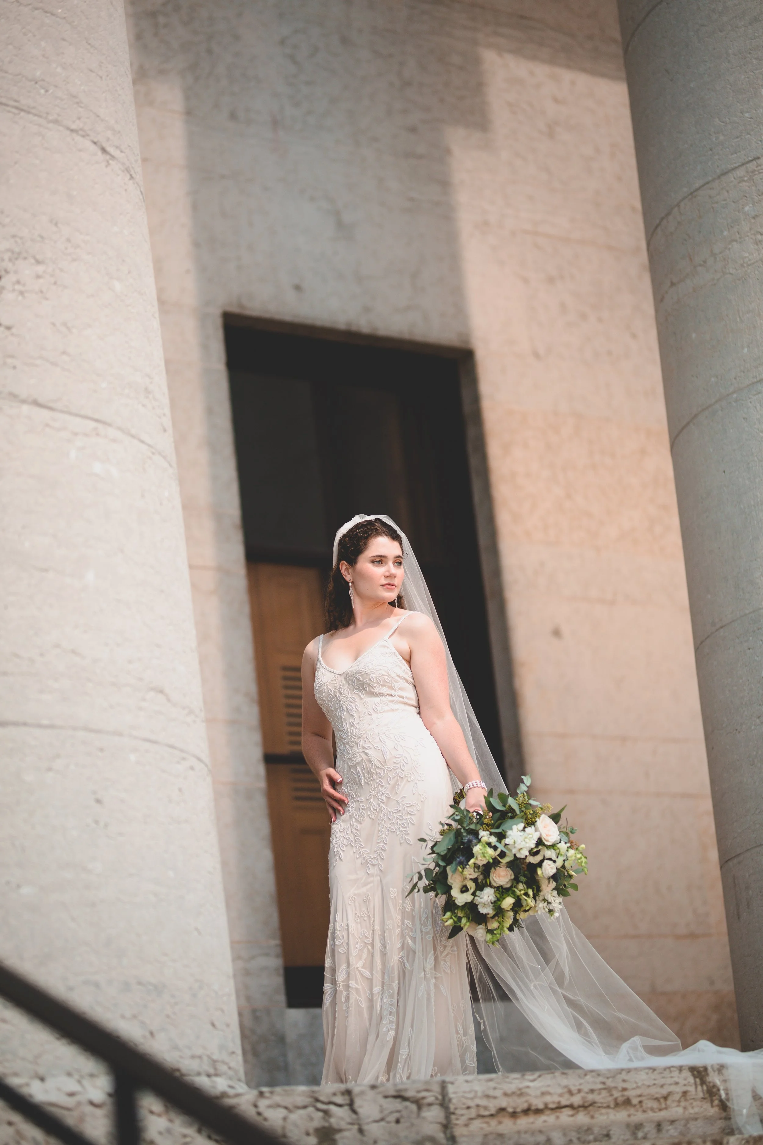 A bride in a white wedding dress with a veil holding a bouquet of white and green flowers standing outside a building with large stone columns.