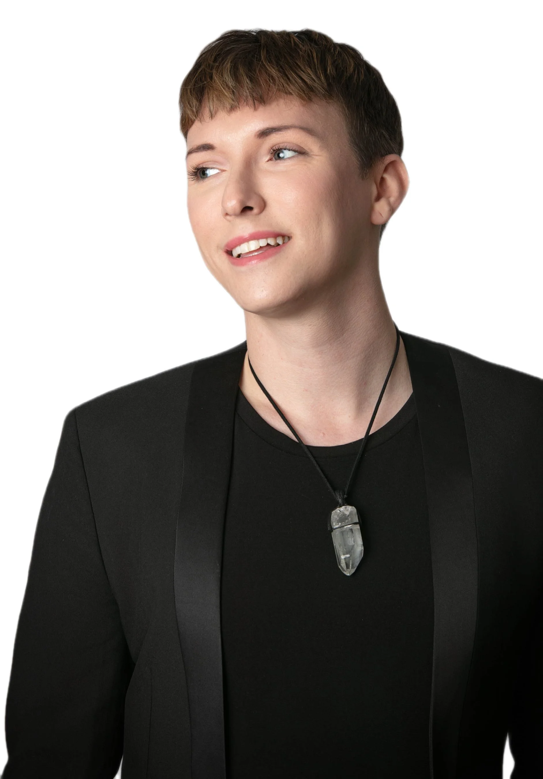 A young woman with short brown hair and blue eyes, wearing a black blazer, black shirt, and a necklace with a clear quartz pendant, smiling slightly against a white background.