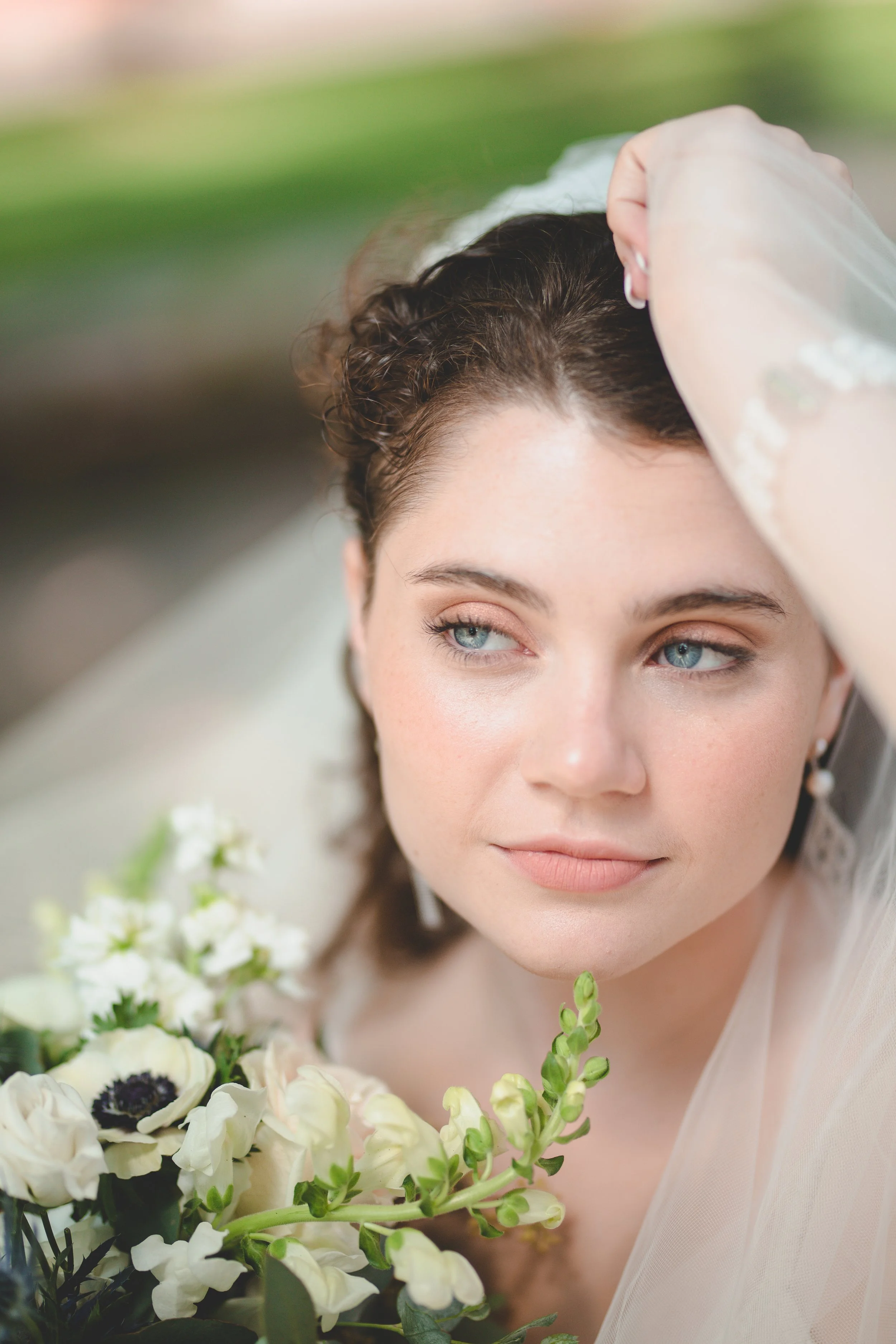 Close-up of a bride with blue eyes, wearing a white wedding gown and veil, holding a bouquet of white and cream flowers, with a blurred green background.