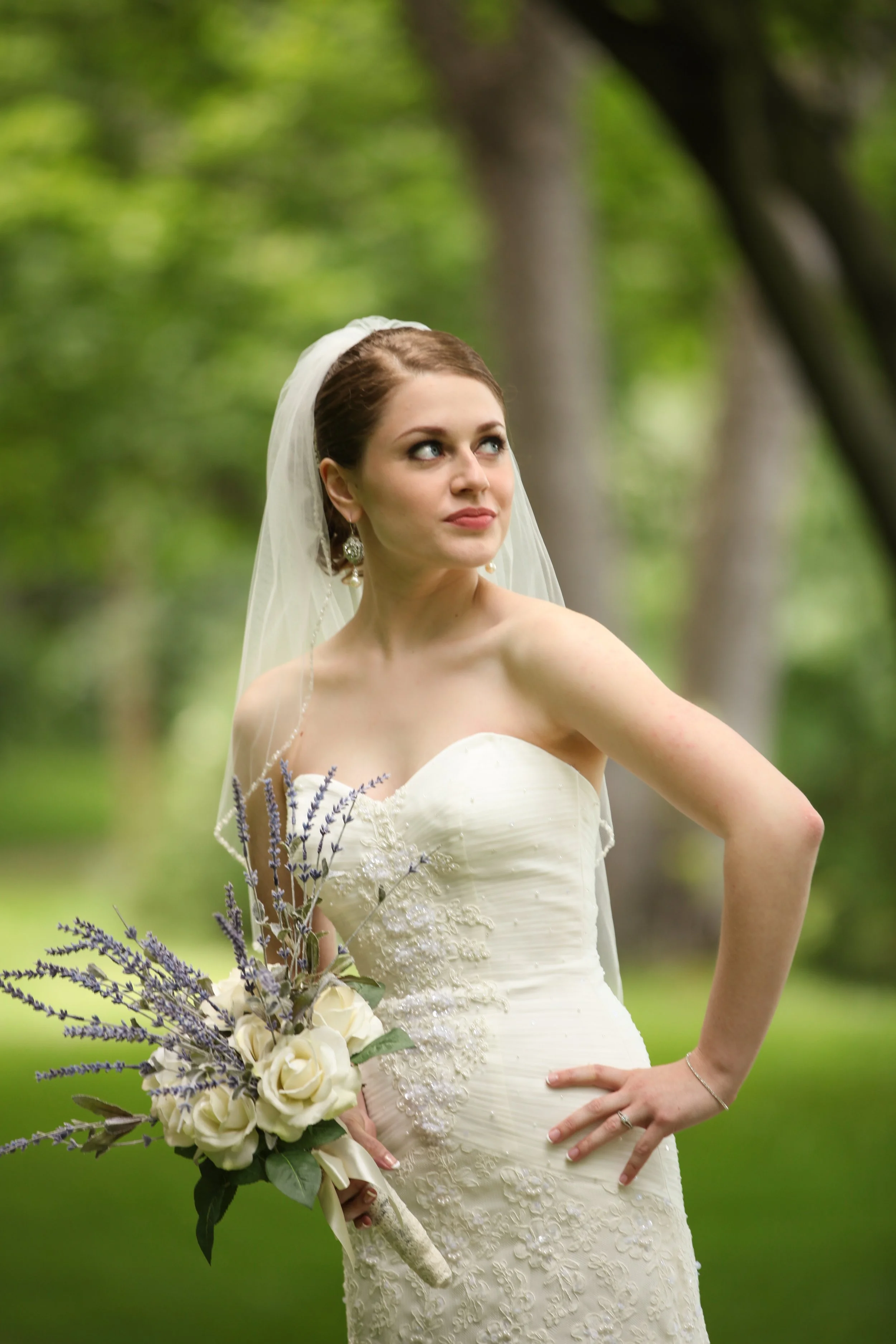 Bride in white wedding dress holding bouquet of white roses and lavender outdoors, looking off to the side.