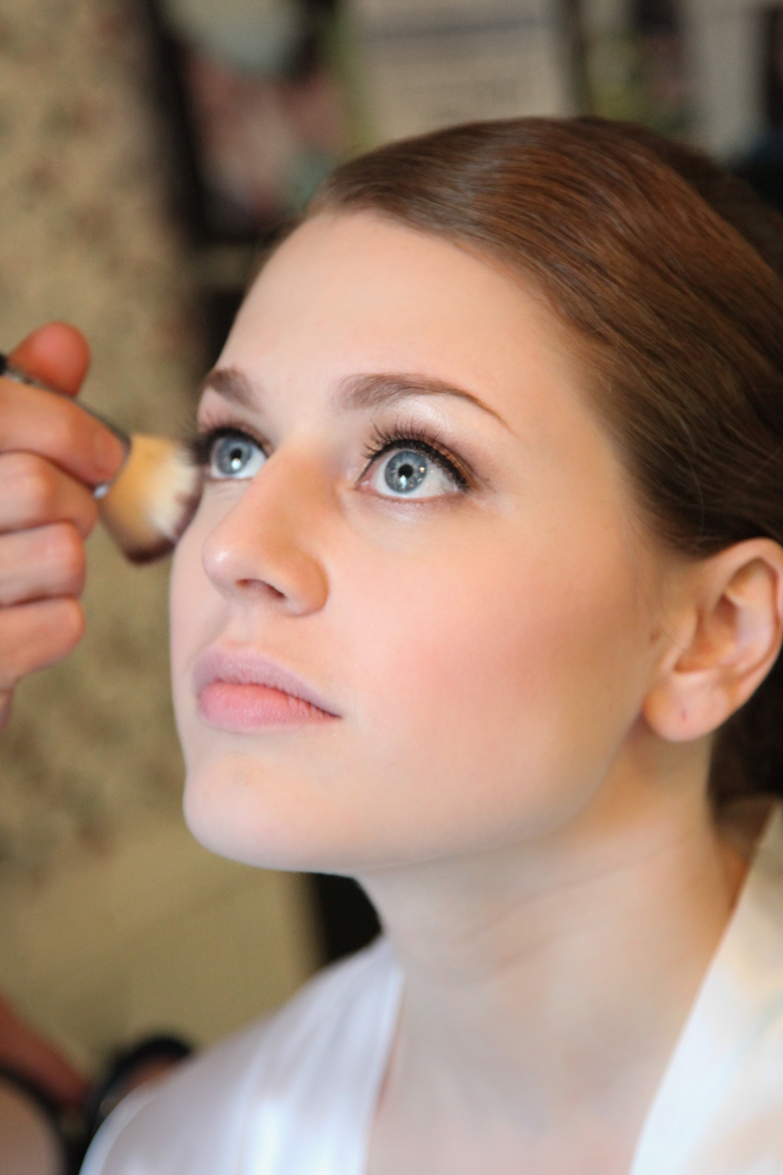 A woman with blue eyes and light makeup having her face touched up with a makeup brush.