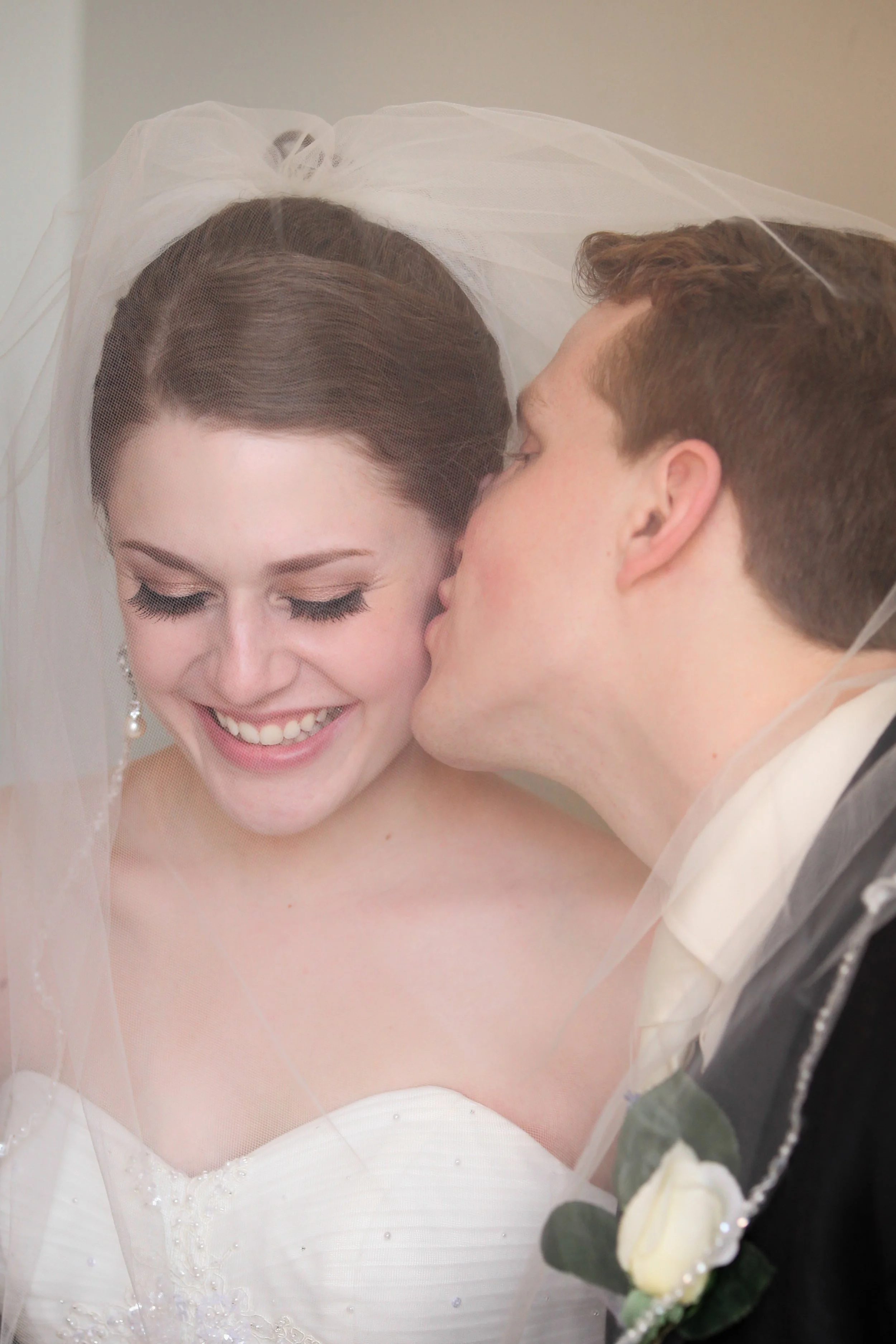 A bride with dark hair and a veil smiling as a groom kisses her cheek.