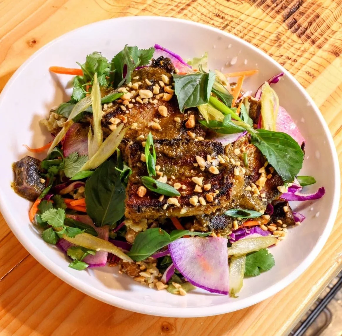 A salad with grilled chicken, mixed greens, chopped nuts, sliced radishes, shredded carrots, and herbs in a white bowl on a wooden table.