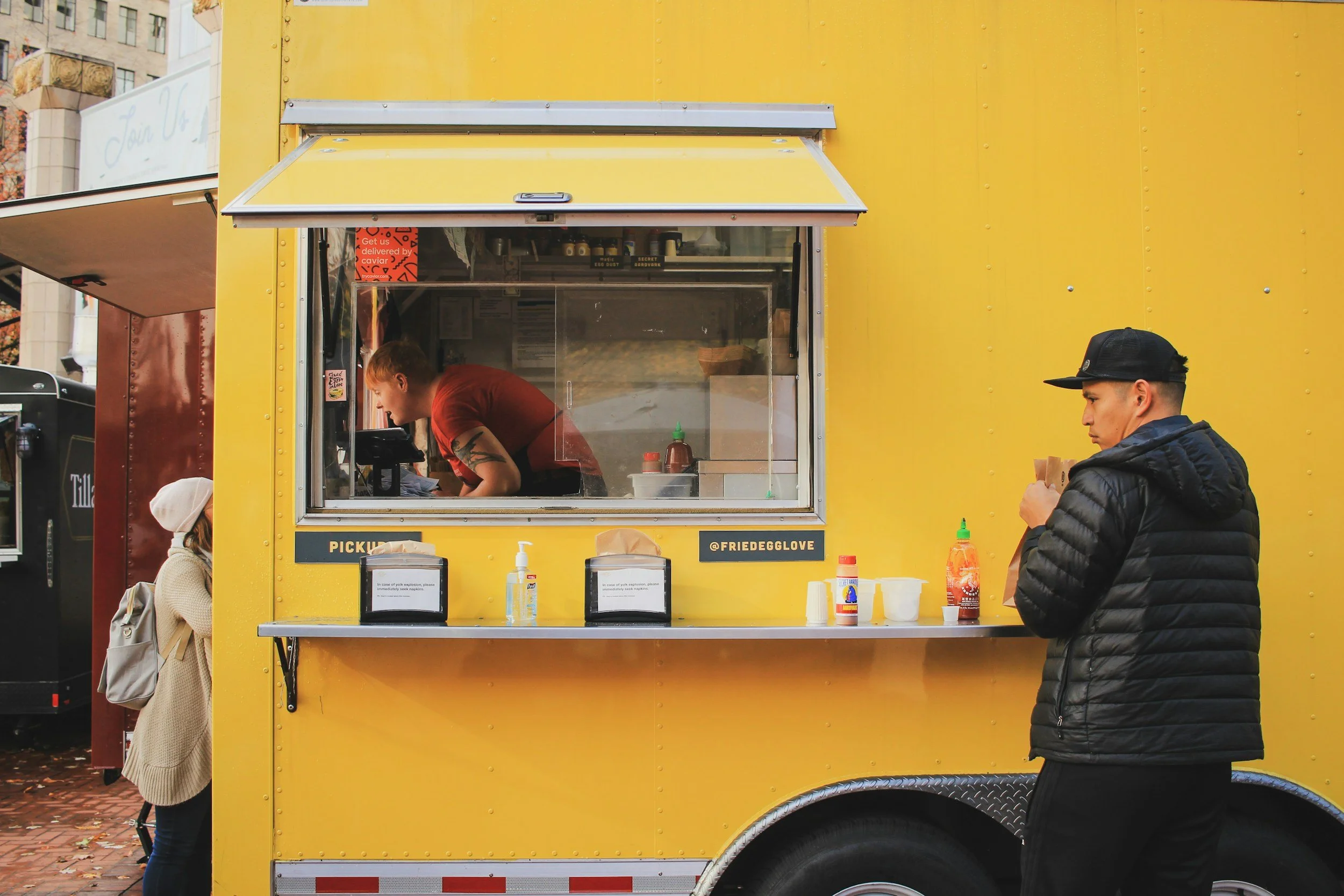 A yellow food truck with a window where a worker is preparing food, and a man in a black jacket and cap ordering food, while a young girl in a beige coat and white beanie waits nearby.