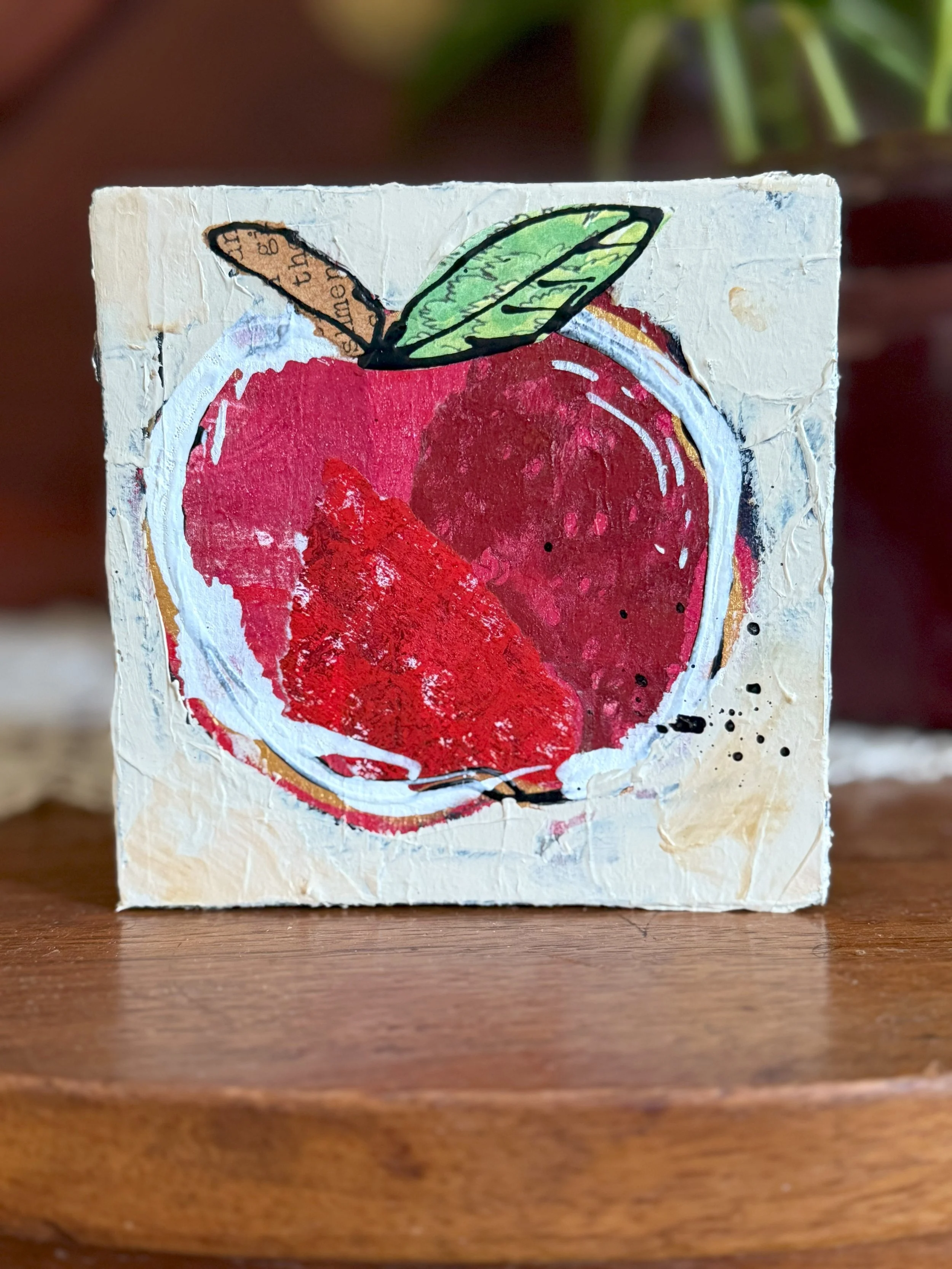 A small square piece of paper or canvas with a colorful, artistic depiction of a red apple with a green leaf on top, in a mixed media style.