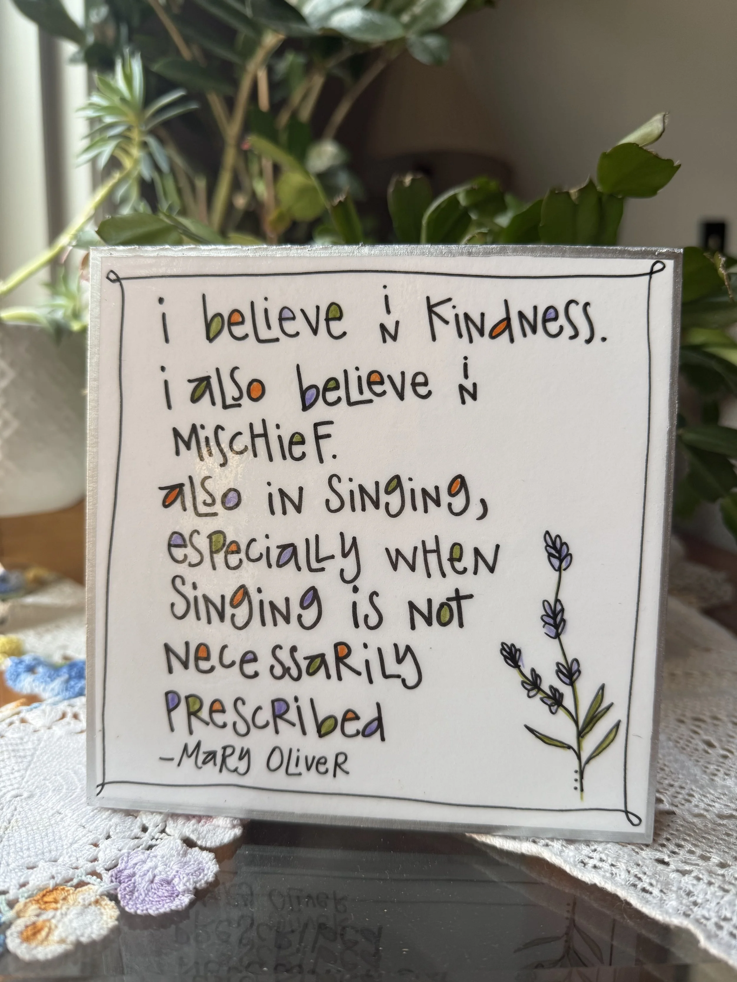 A whiteboard with a handwritten quote by Mary Oliver, decorated with colorful letters, drawings of lavender and flowers, placed on a lace cloth with plants in the background.