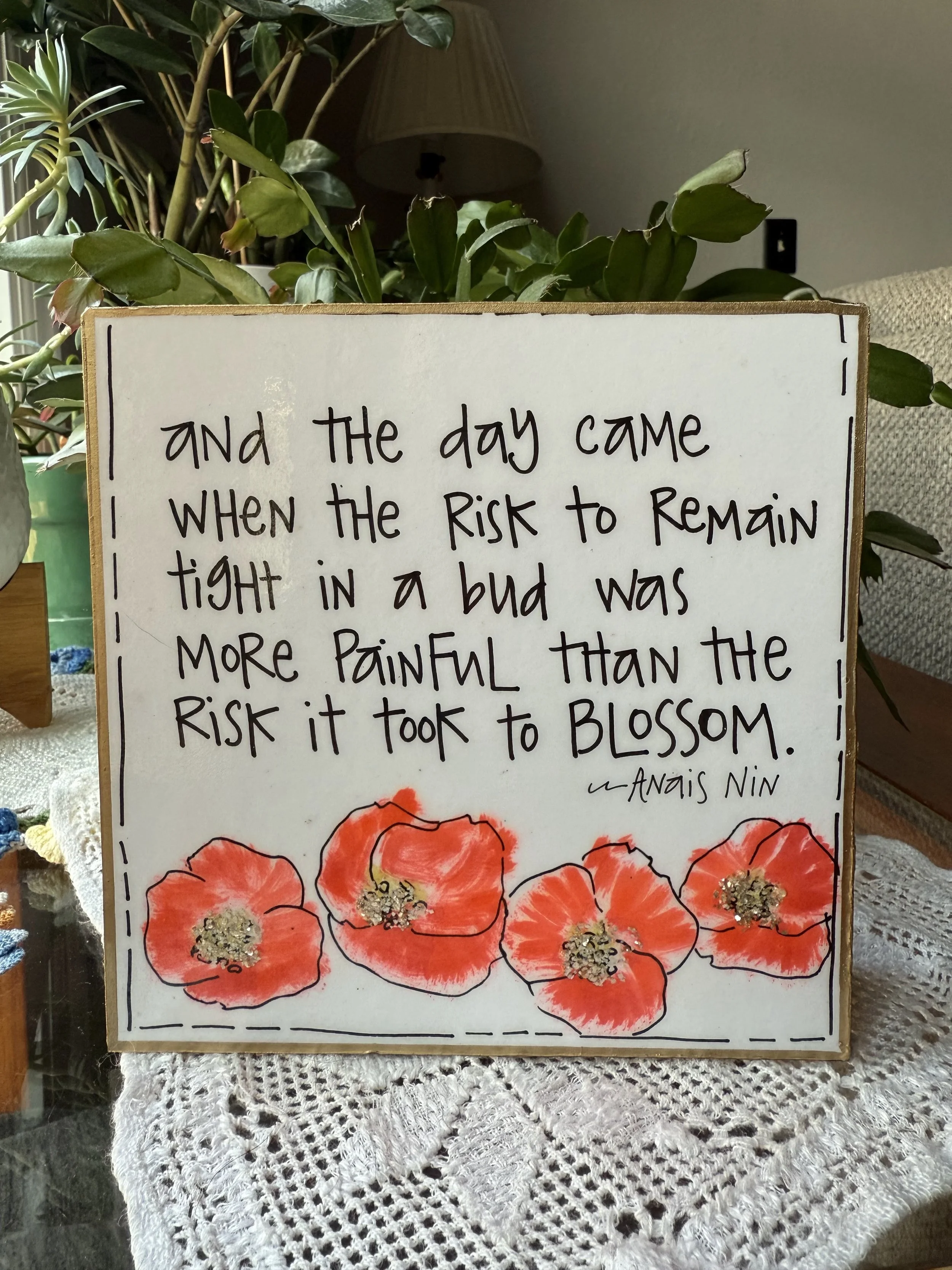 A decorative sign with a handwritten quote by Anaïs Nin, surrounded by orange-red poppies illustrations, placed on a lace tablecloth near a leafy houseplant.