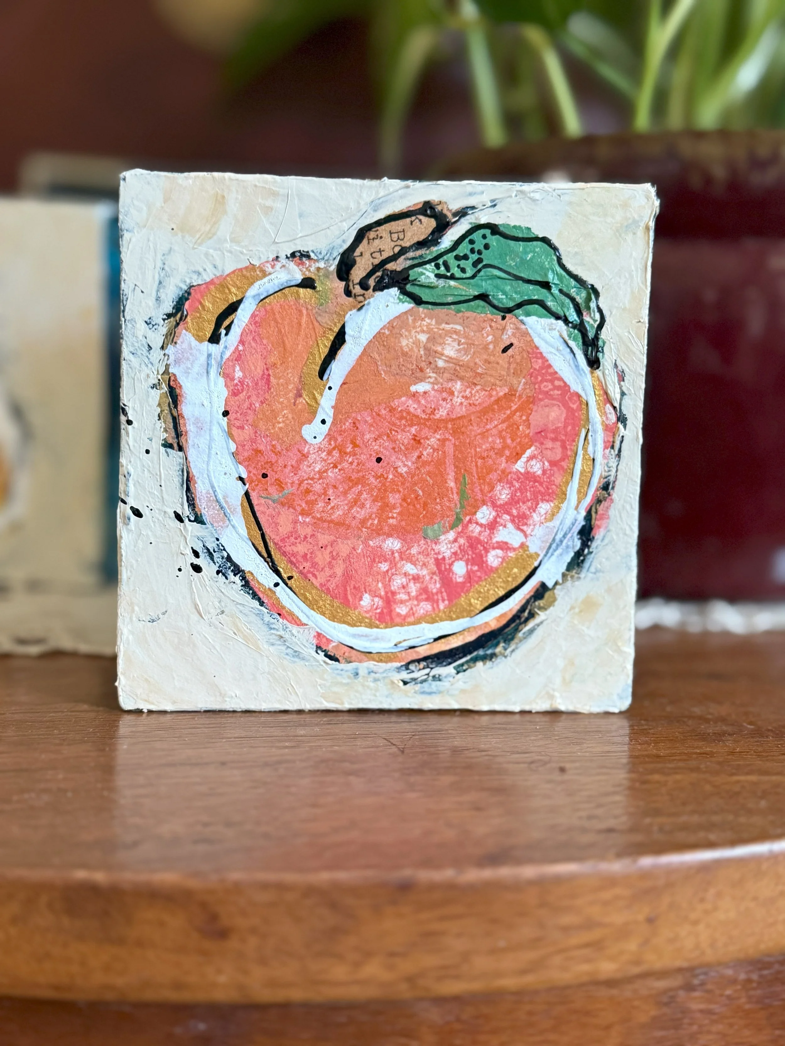 A small, textured canvas painting of a peach with a green leaf, created with bold, abstract brushstrokes and outlines, placed on a wooden surface with blurred plants in the background.