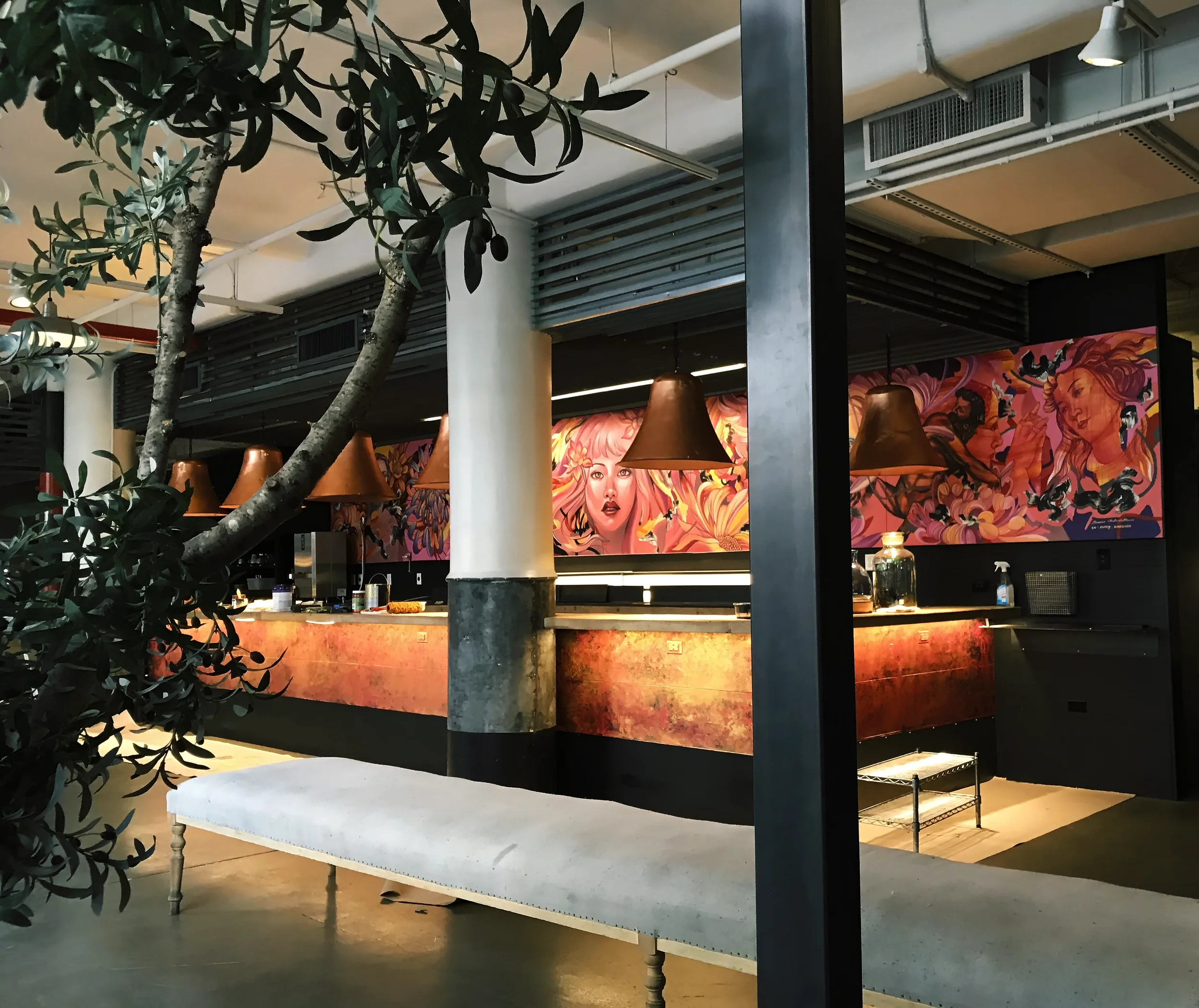 Indoor area with a partial view of a display featuring colorful artwork of a woman with long hair, multiple hanging pendant lights, part of a tree in the foreground, and industrial-style ceiling vents and lighting.