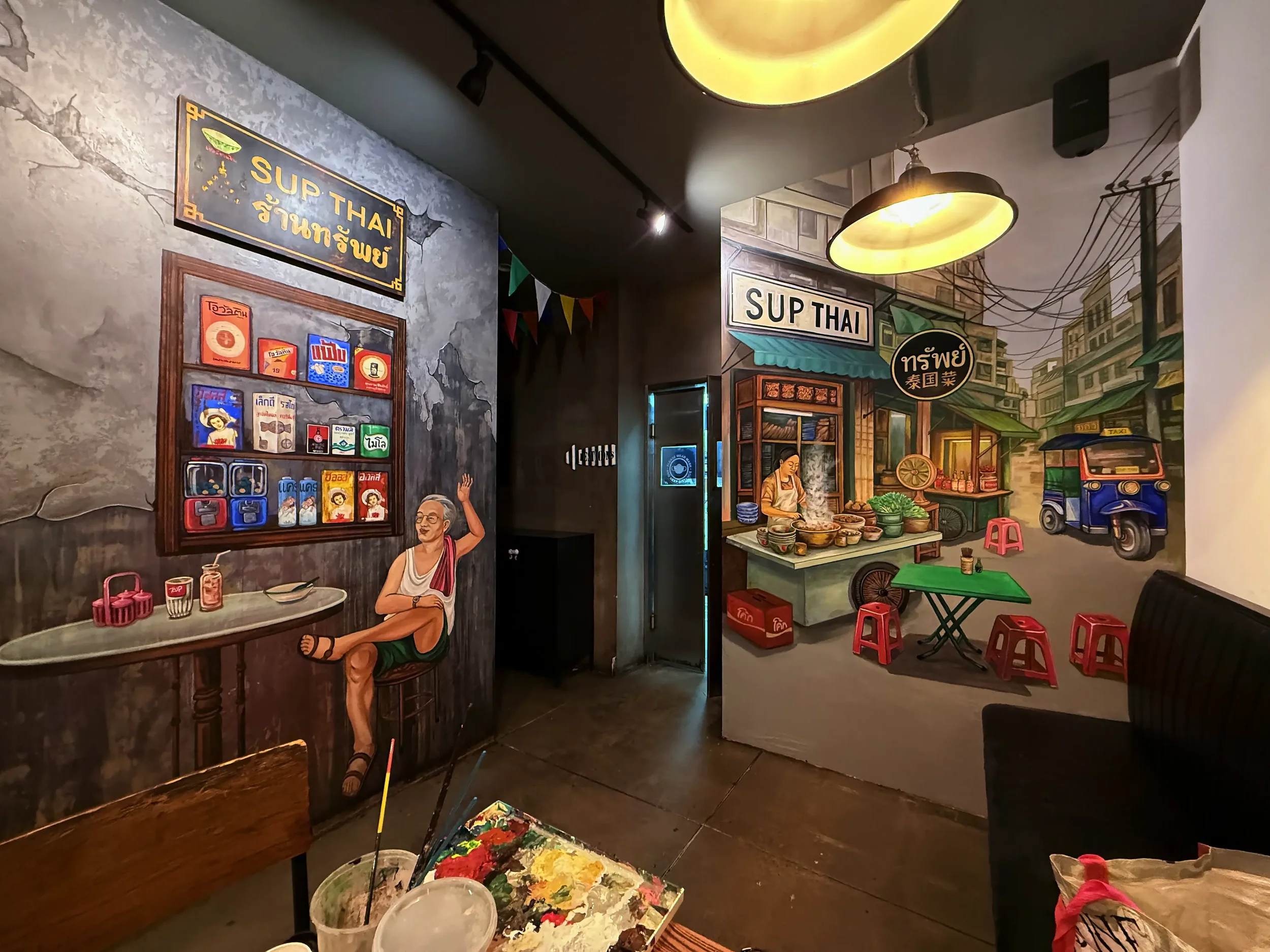 Decorative mural of a street scene with a woman shopkeeper, small tables with red stools, and a mural of a cityscape with buildings, trees, and a tuk-tuk vehicle, with hanging lights.