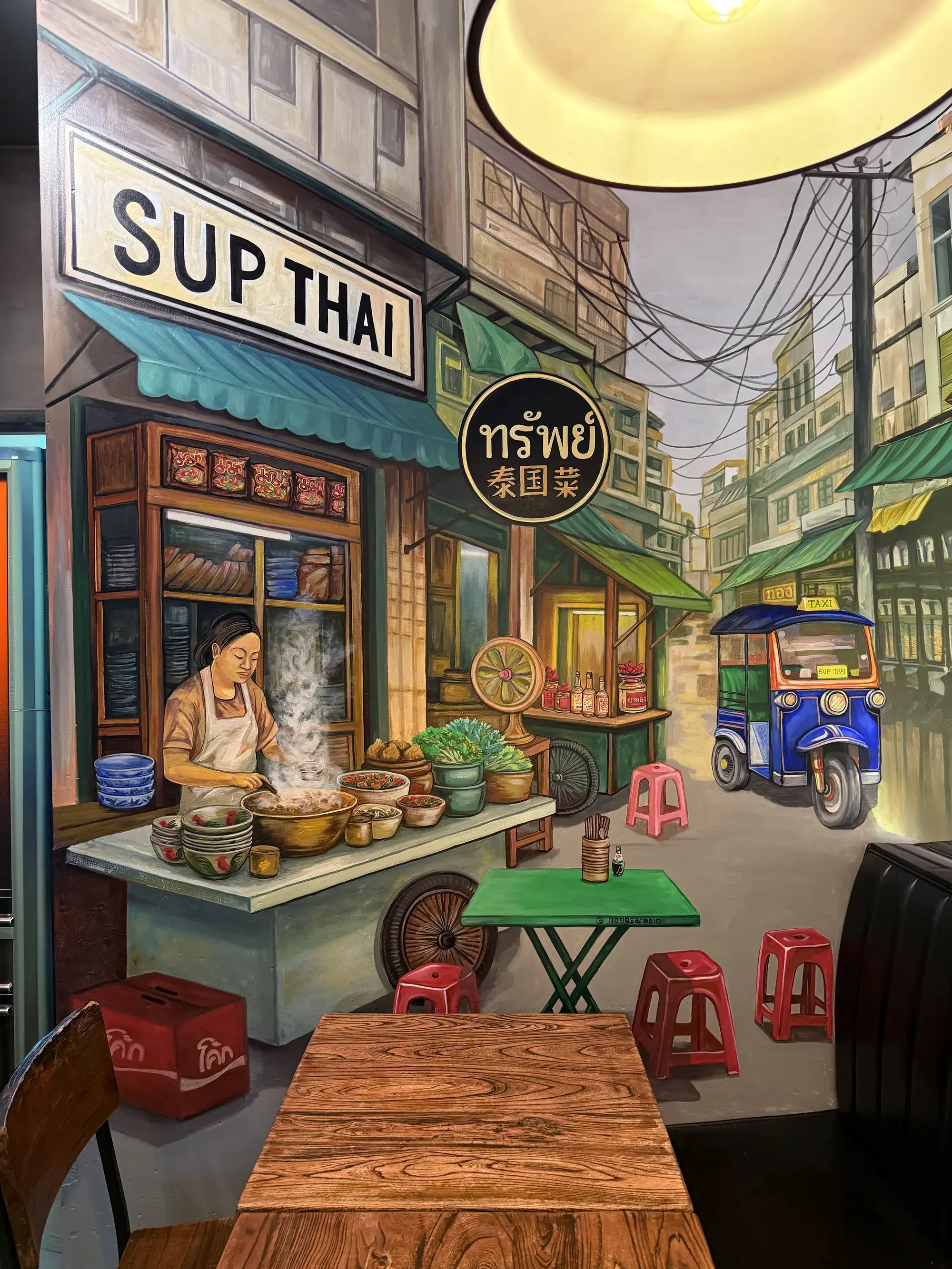 A mural of a street scene featuring a woman preparing food at a street stall with signs in English and Thai, a tuk-tuk vehicle, and small red stools, set in an urban area with buildings and power lines overhead.