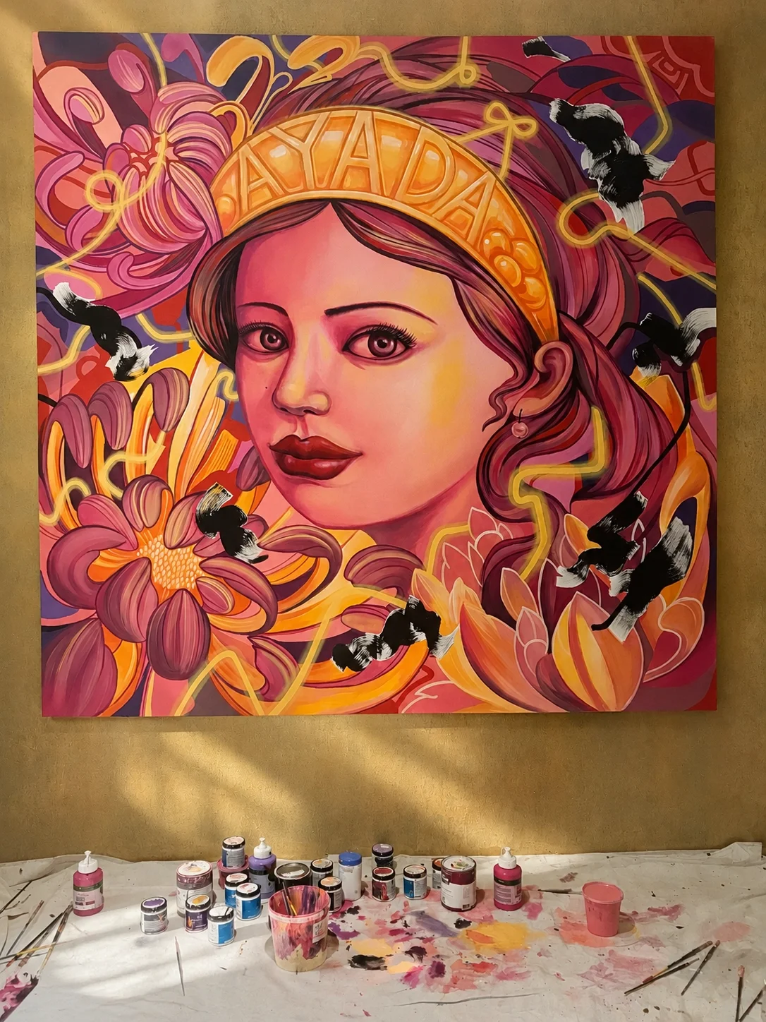 A colorful painting of a woman with pink and purple hair, wearing a gold crown, surrounded by pink flowers and black and white birds, with painting supplies below.