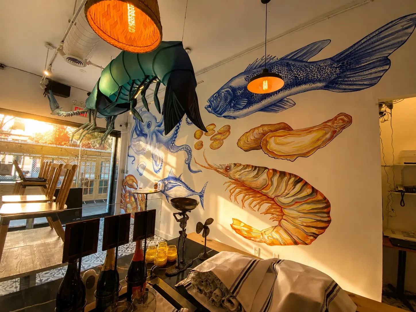 Colorful ocean-themed mural with a large fish, shrimp, an octopus, and an insect hanging from the ceiling in a restaurant interior, with sunlight coming through the window.