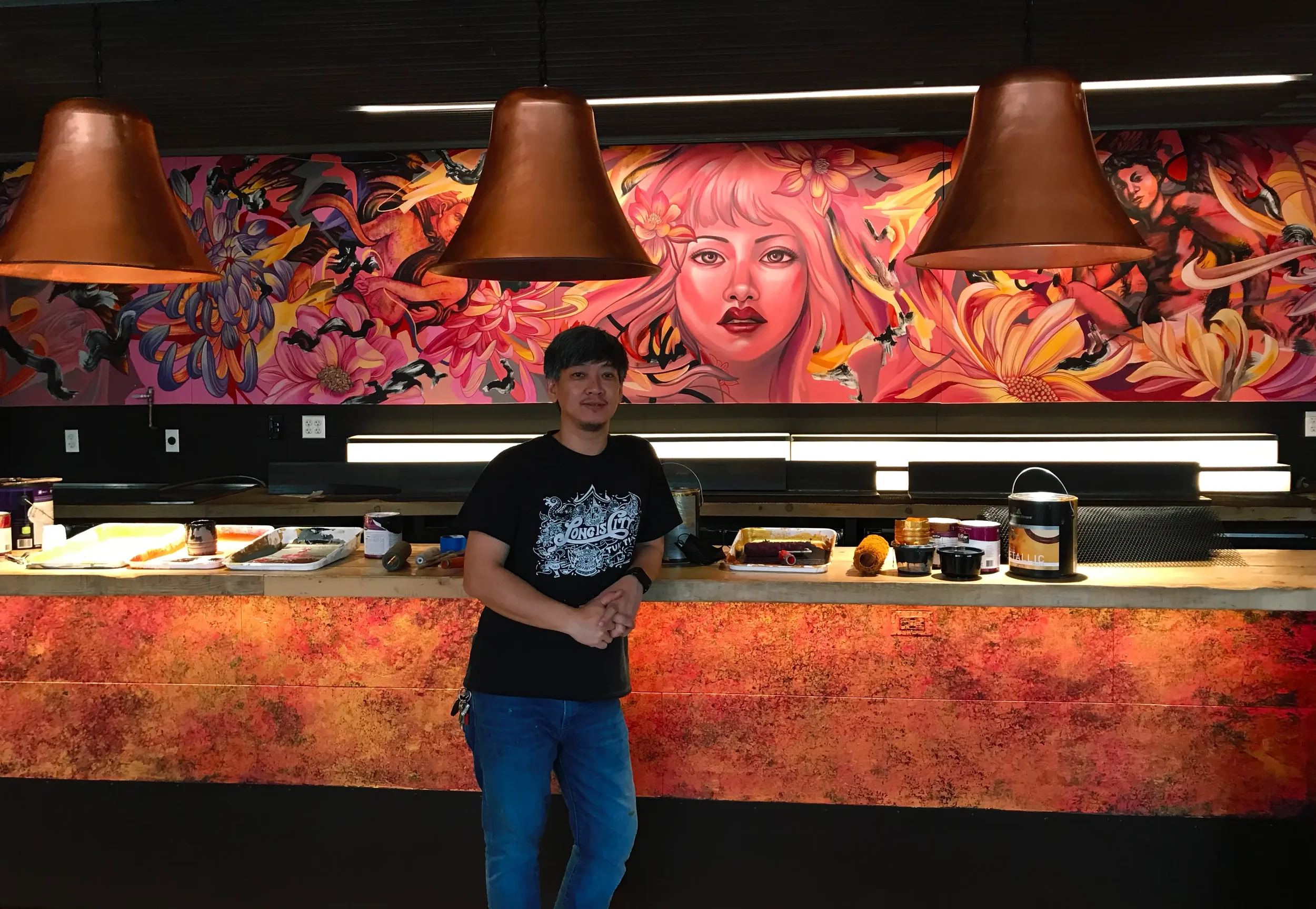A man with dark hair and a mustache, wearing a black t-shirt, standing in front of a colorful mural of a woman with pink hair, surrounded by large flowers and abstract designs. The area appears to be a modern indoor space with hanging lights and a counter with various items.