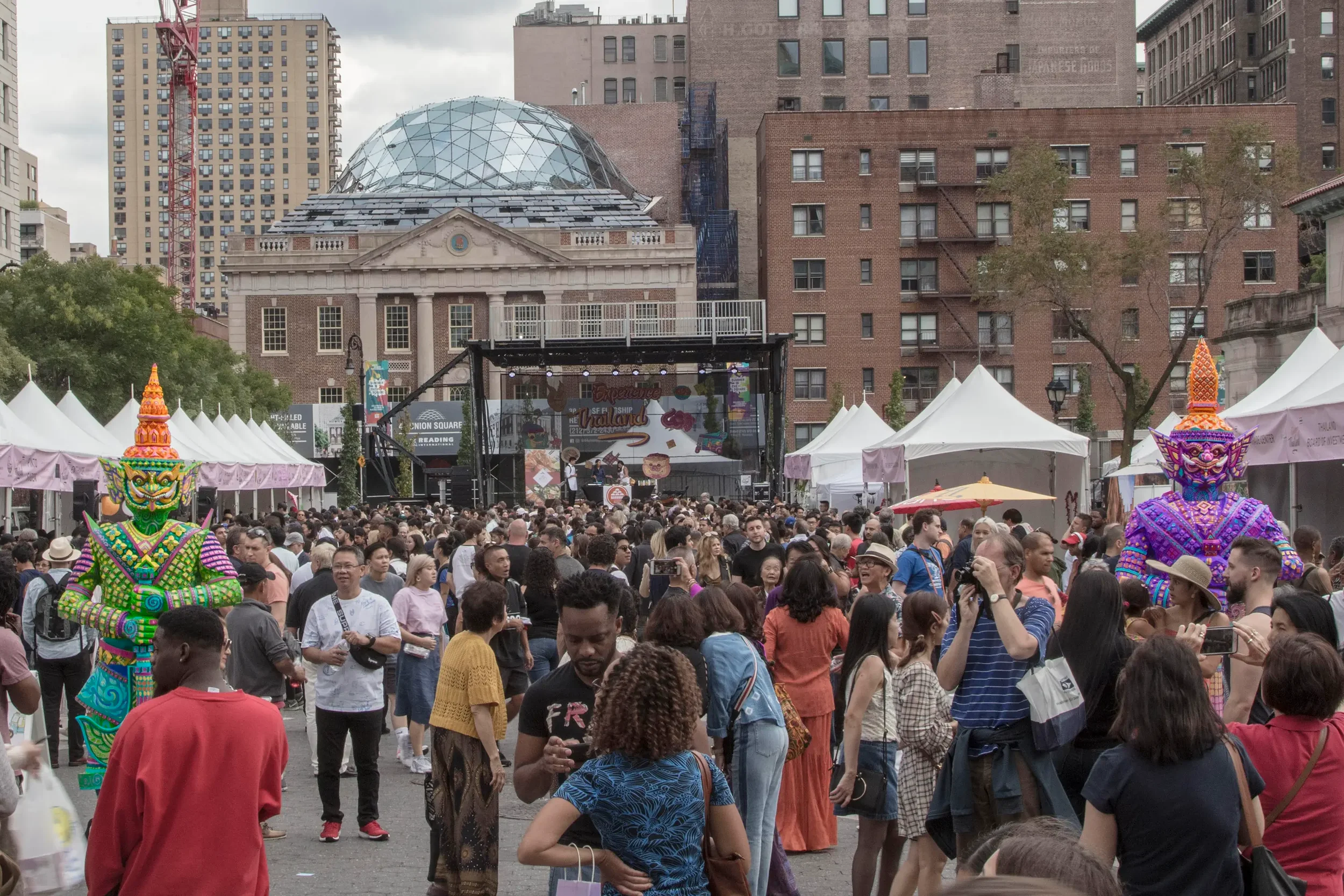 A large crowd of people gathered outdoors at a festival or event. There are tents with Asian-themed decorations, including a vibrant, colorful figure in traditional costume in the foreground. In the background, there are high-rise buildings and a stage with a banner that says they are at Union Square.