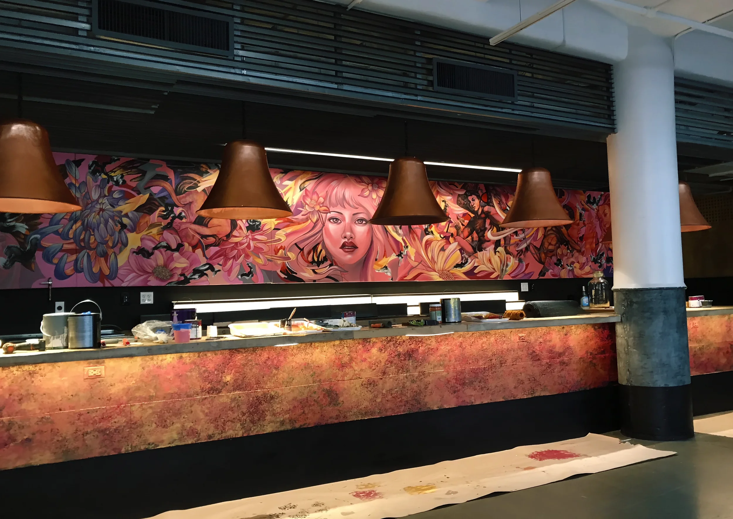 Interior of a building with a colorful mural of a woman's face and flowers, brown hanging lamps, a marble counter with art supplies, and a white column on the right side.