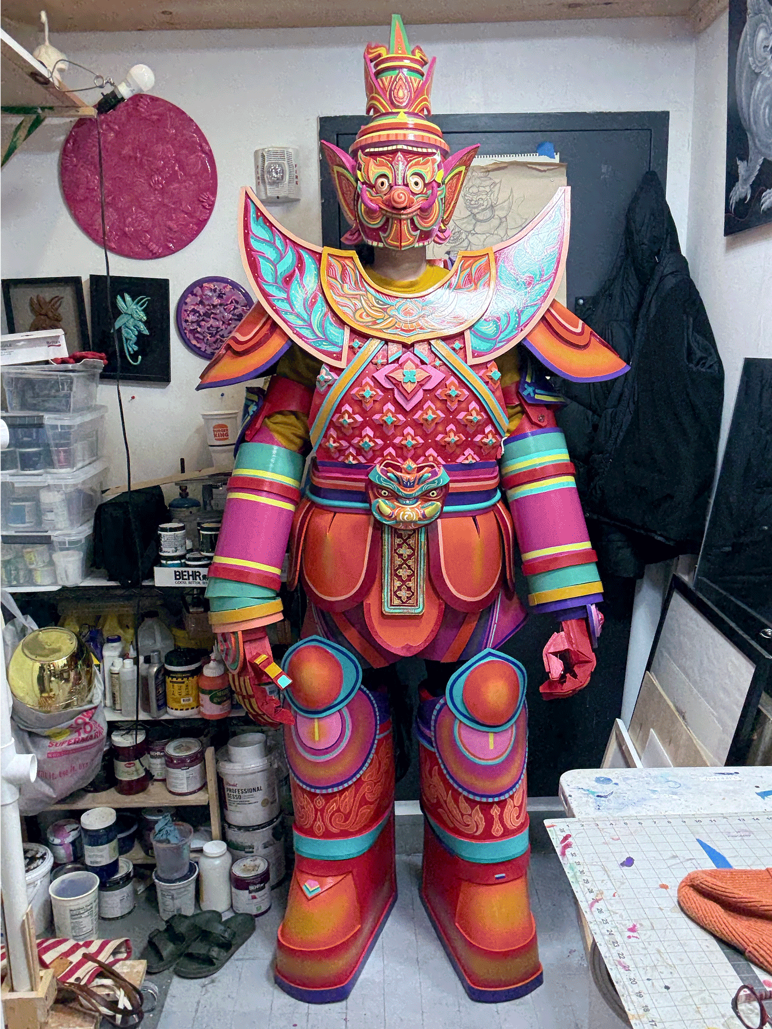 A person dressed in a colorful, elaborate costume resembling a traditional or mythical figure, standing in a workshop among art supplies and paintings. The costume features a large, detailed mask with exaggerated facial features and a crown-like headdress, along with intricate patterns and vibrant colors.