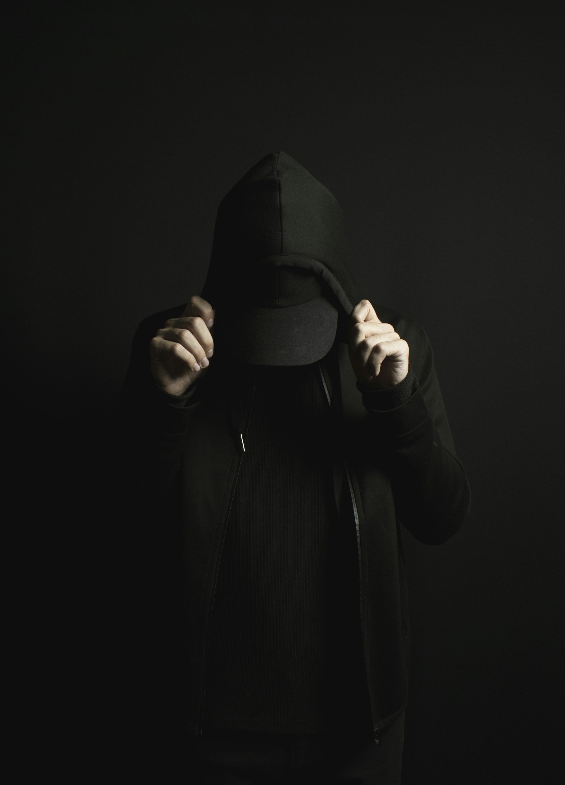 Person wearing a black hoodie and cap, holding the hood with both hands, against a dark background.