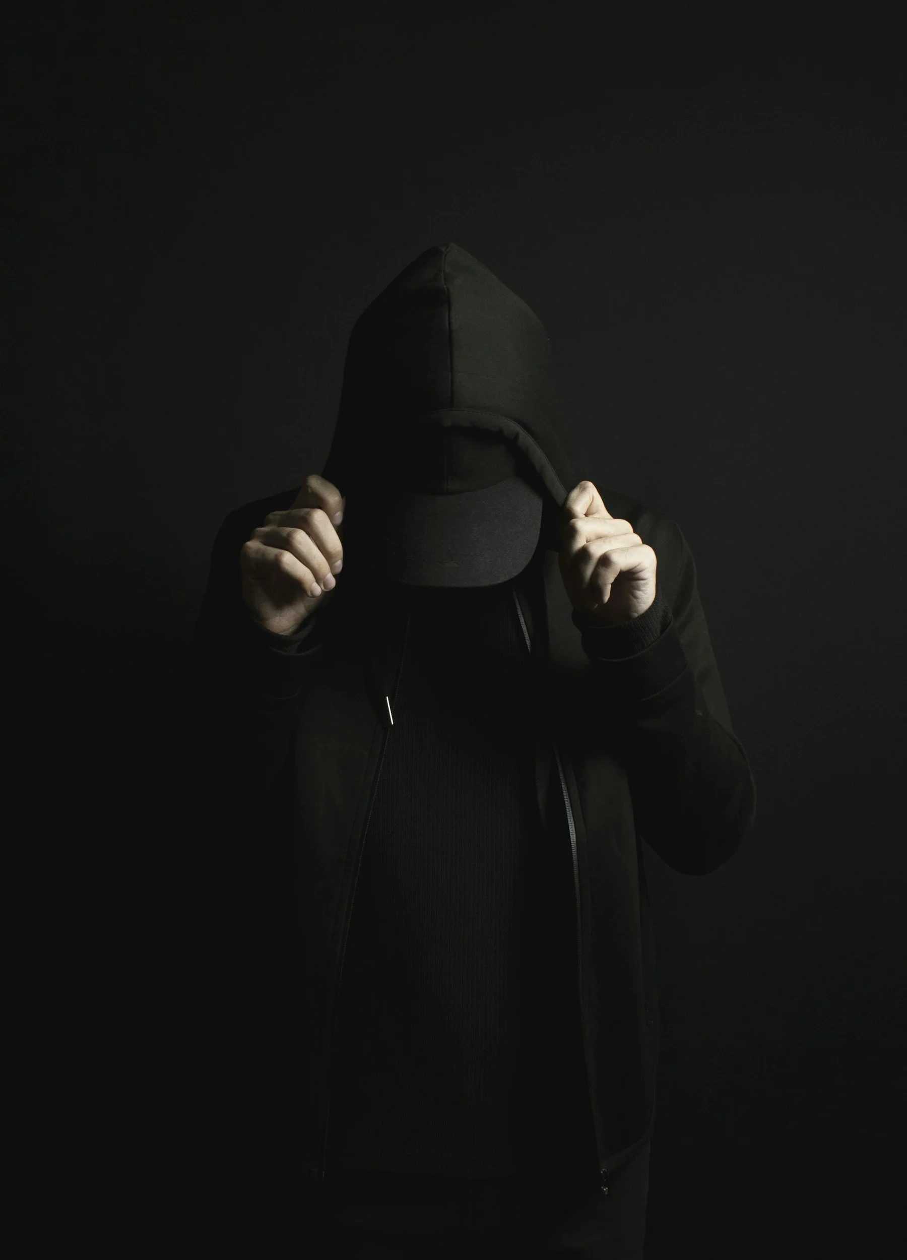 Person wearing a black hoodie and cap, hiding their face, holding the hoodie with both hands, against a dark background.