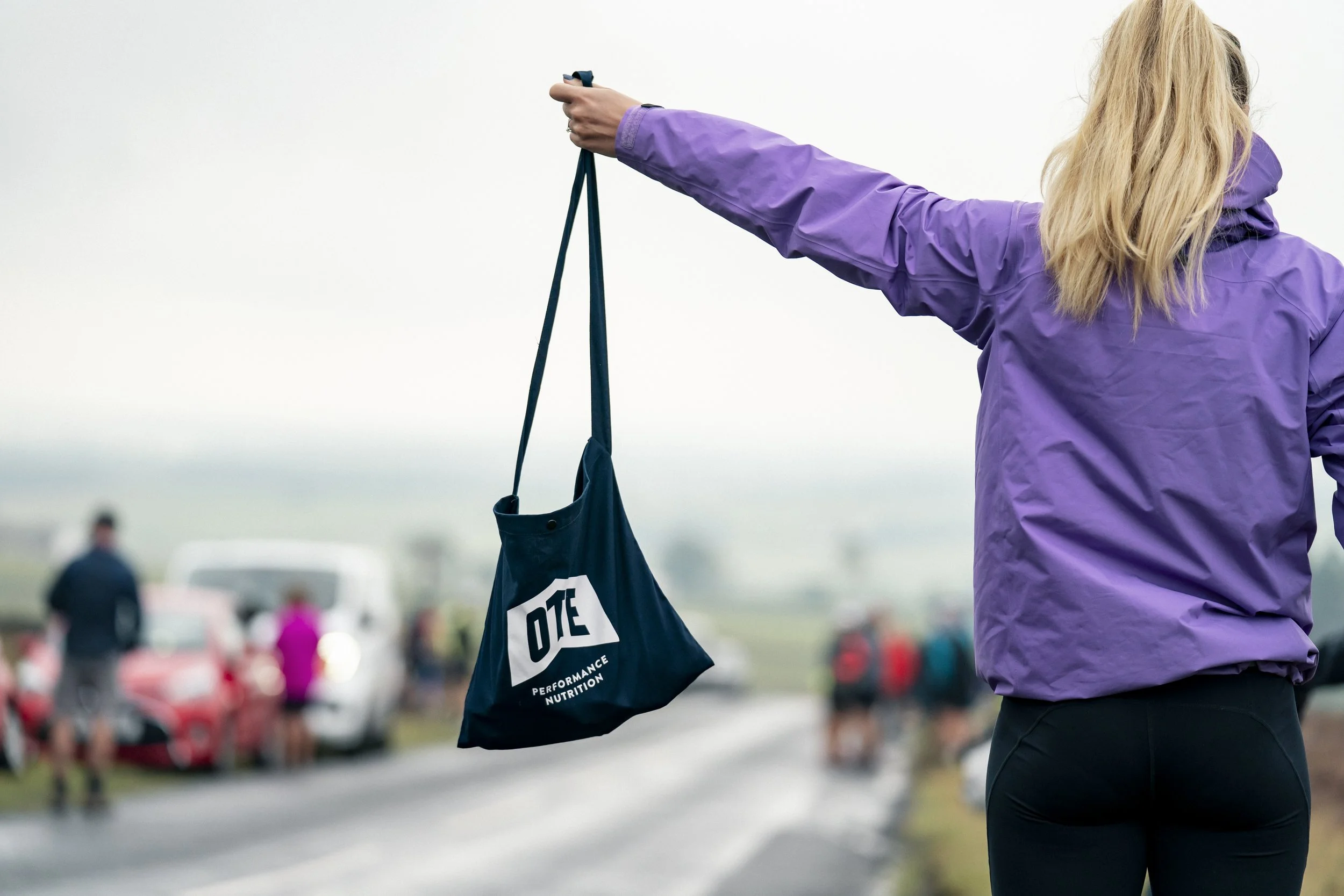 A woman in a purple jacket holds a black bag with white text that says "DITE Performance Nutrition" while standing on a road with other people and cars in the background, outdoors on a cloudy day.