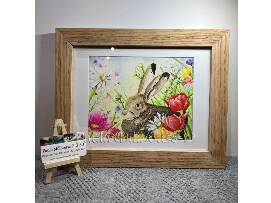 Brown hare sitting among colorful spring wildflowers including tulips and daisies, wildlife watercolor by Paula Millhouse.