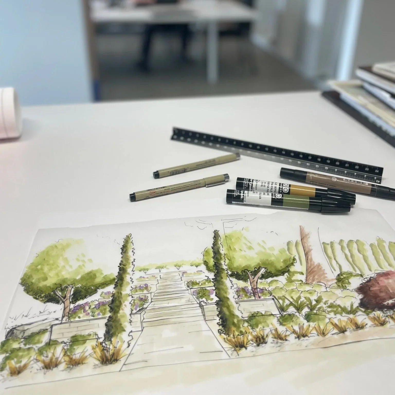 A rendered watercolor or ink drawing of a landscaped garden with trees, shrubs, and steps leading to a pathway, depicted on a piece of paper on a white table, with drawing pens and markers around it.