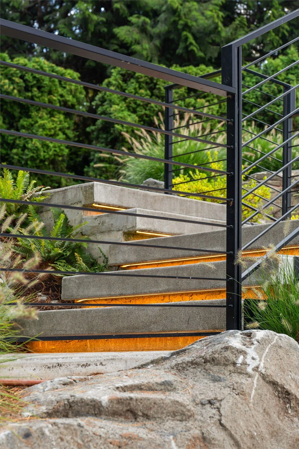 Modern outdoor staircase with concrete steps and black metal railing surrounded by greenery and rocks.