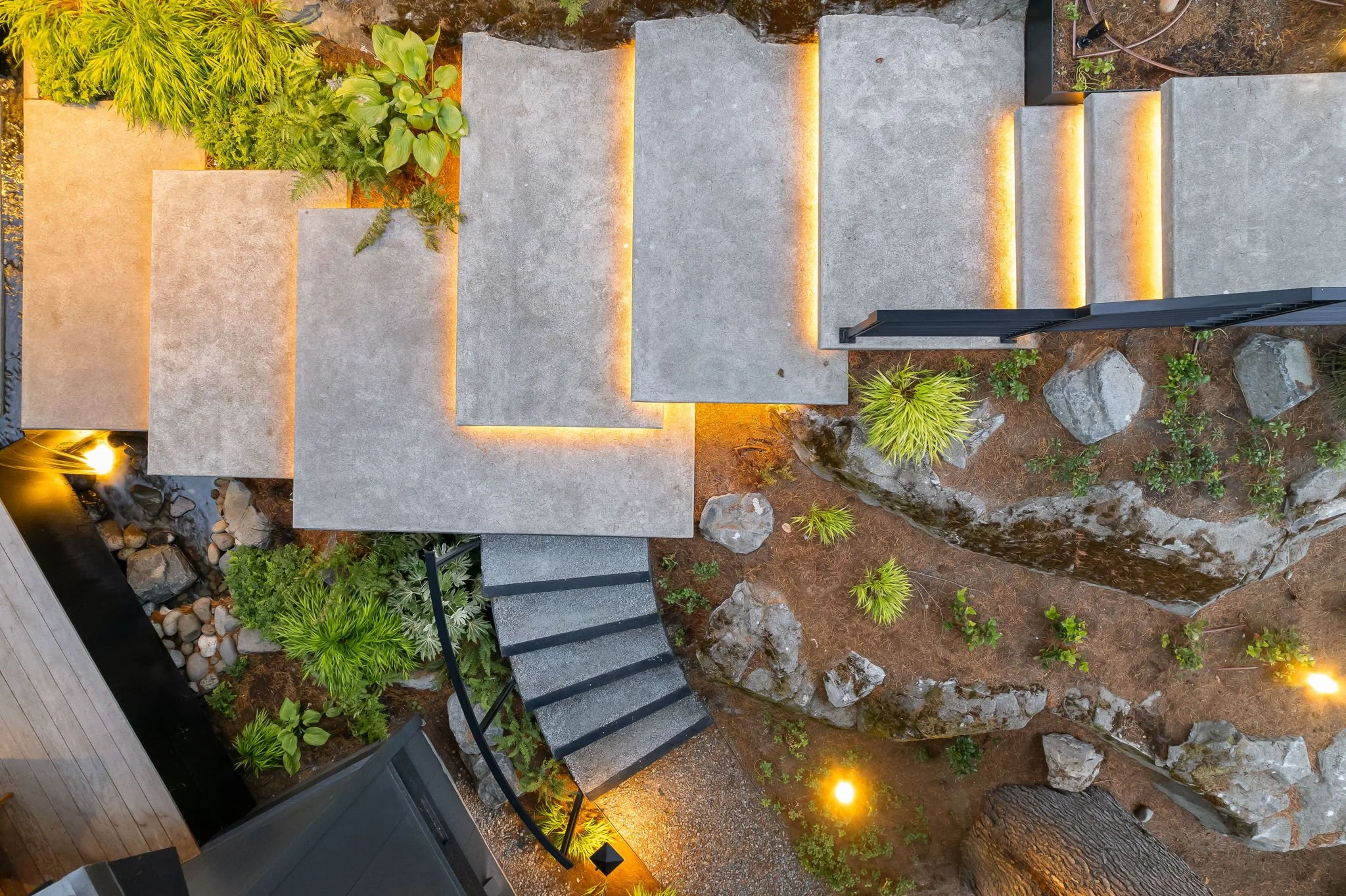 Aerial view of a modern outdoor landscape with concrete steps, surrounding plants, rocks, small waterfall, and warm lighting.