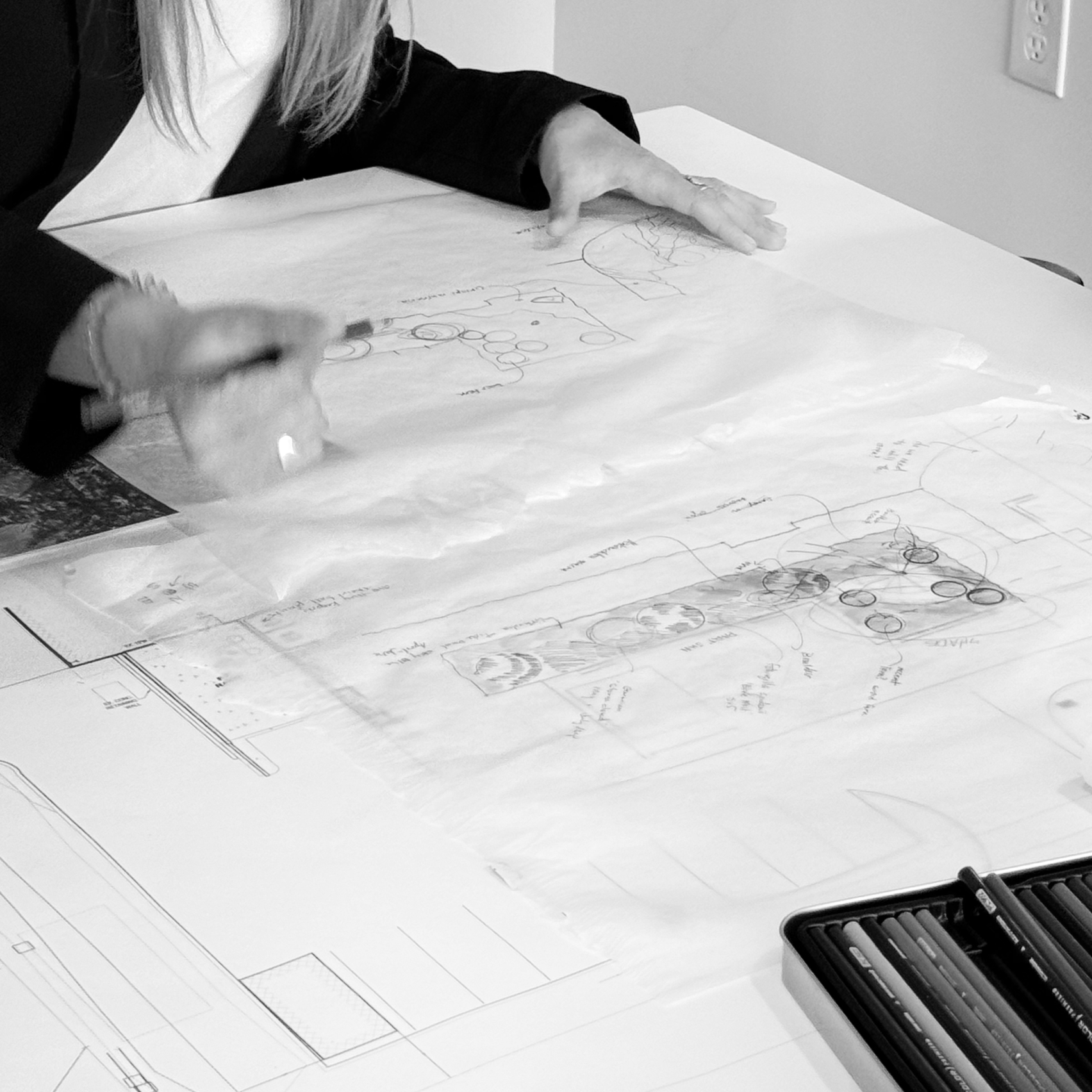 A person working on a large sheet of paper with architectural drawings, using a black pen. There is a set of markers or colored pencils nearby, and a partially visible power outlet on the wall.