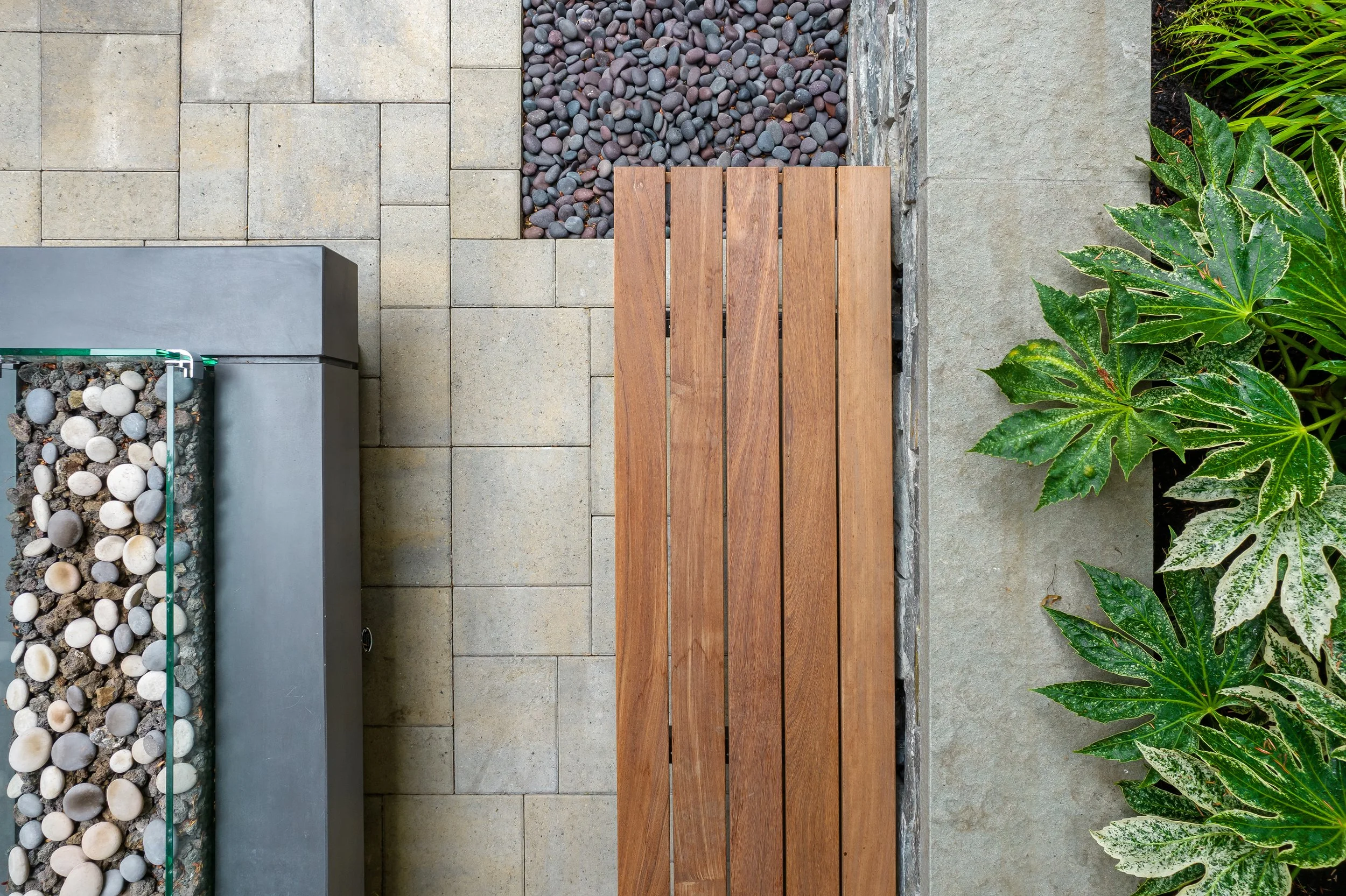 View of an outdoor space with paved stones, two rectangular metal planters with round stones, a wooden bench, and greenery including large variegated and green plants.