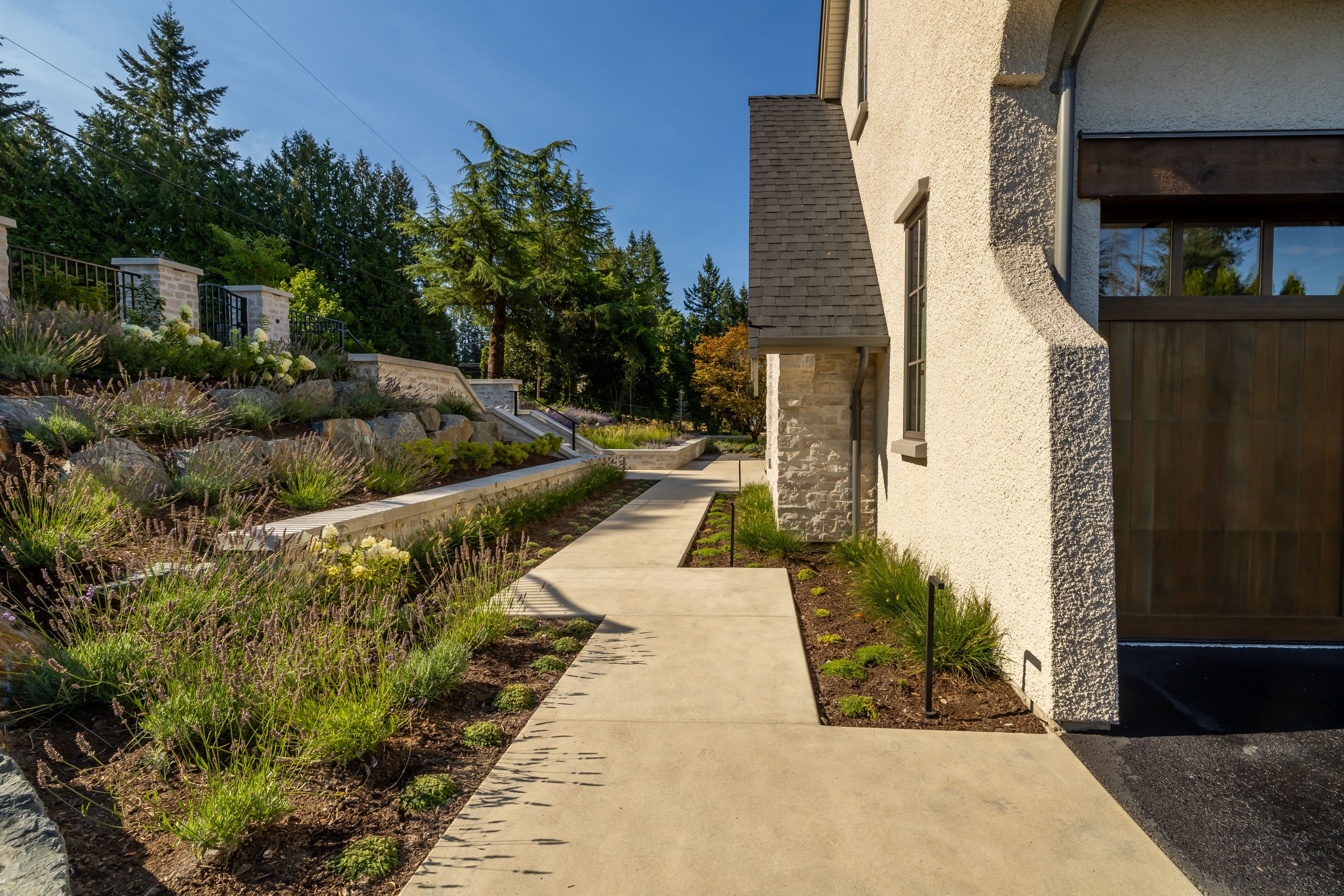 A house with a white textured exterior and a dark brown wooden gate, next to a landscaped yard with flowering plants, trees, and a concrete walkway under a clear blue sky.