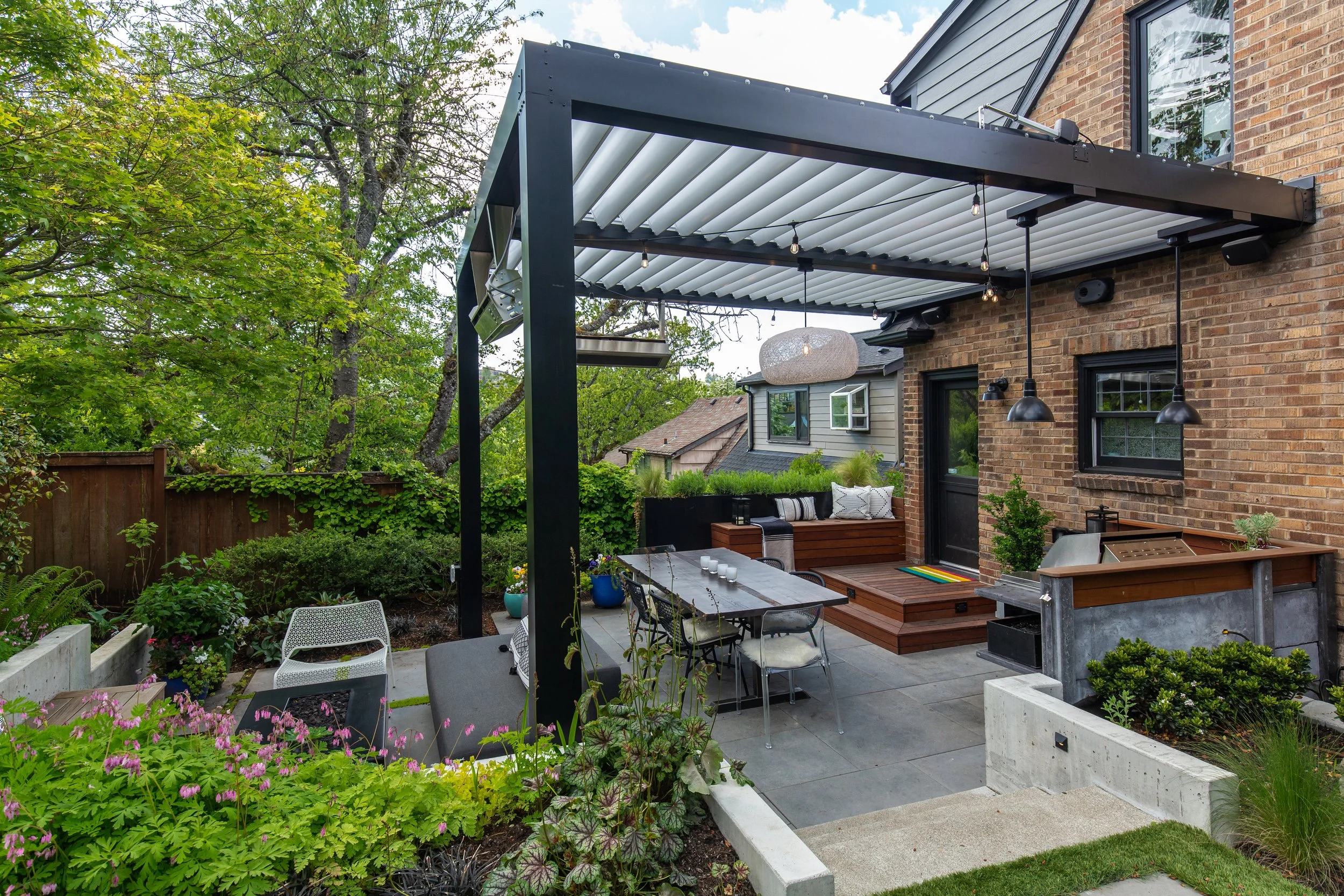 Backyard patio with outdoor dining area, covered by a black metal pergola with fluorescent lights. There are comfy cushions on a built-in bench, surrounded by lush green plants and a brick house with black window frames.