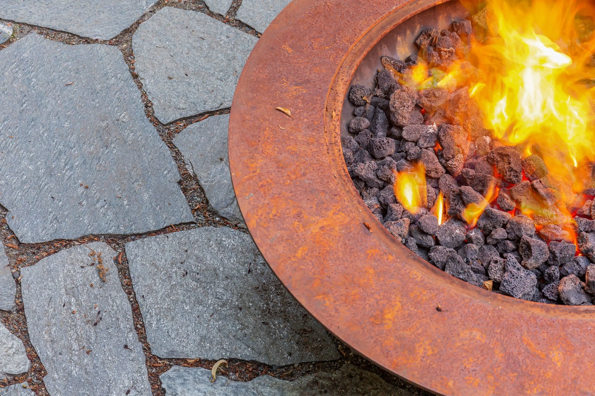 A close-up of a fire pit with glowing flames and charcoal, surrounded by gray stone paving.