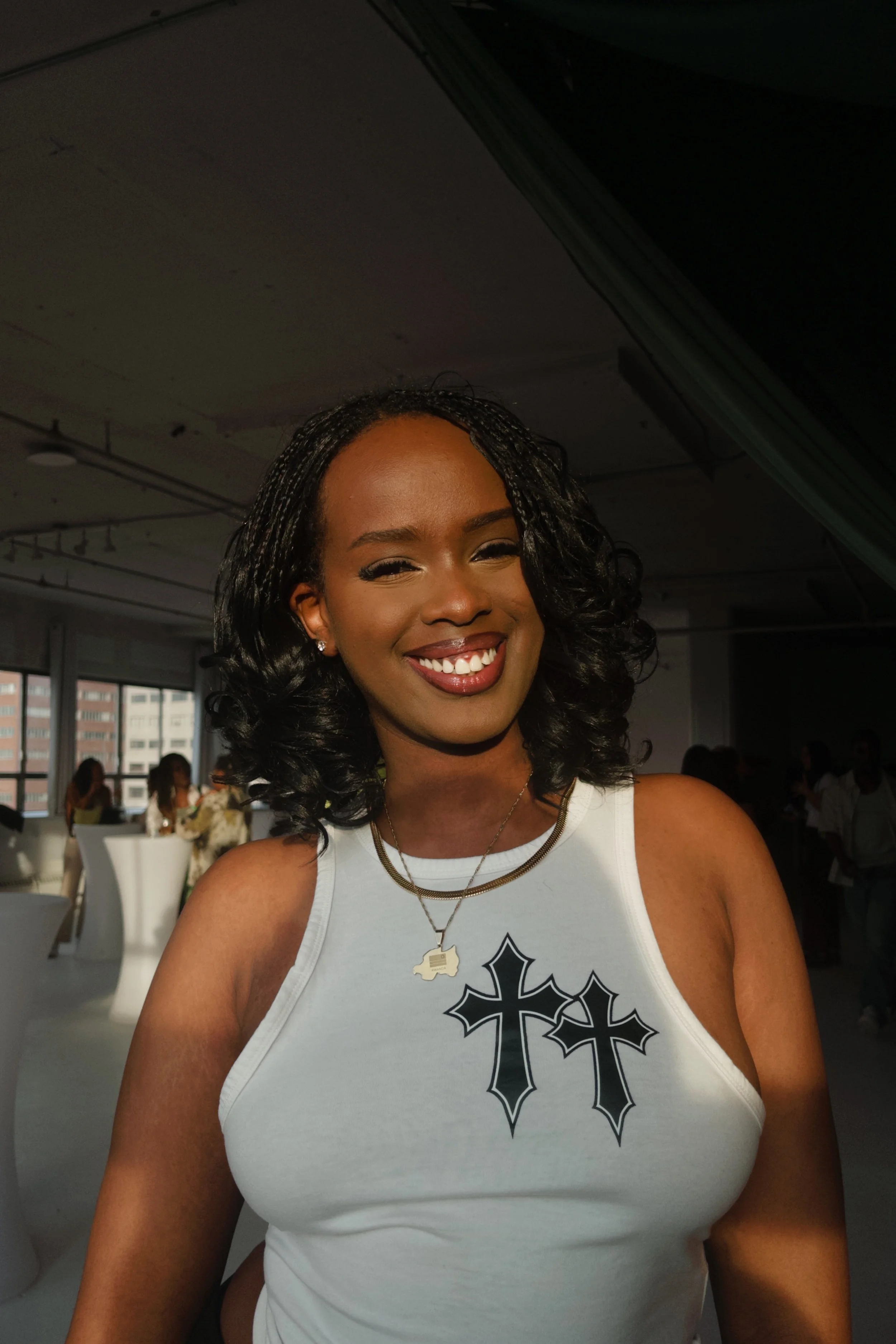 Smiling woman with curly black hair wearing a white sleeveless top with black cross designs, gold necklaces, and earrings at an indoor event with people in the background.