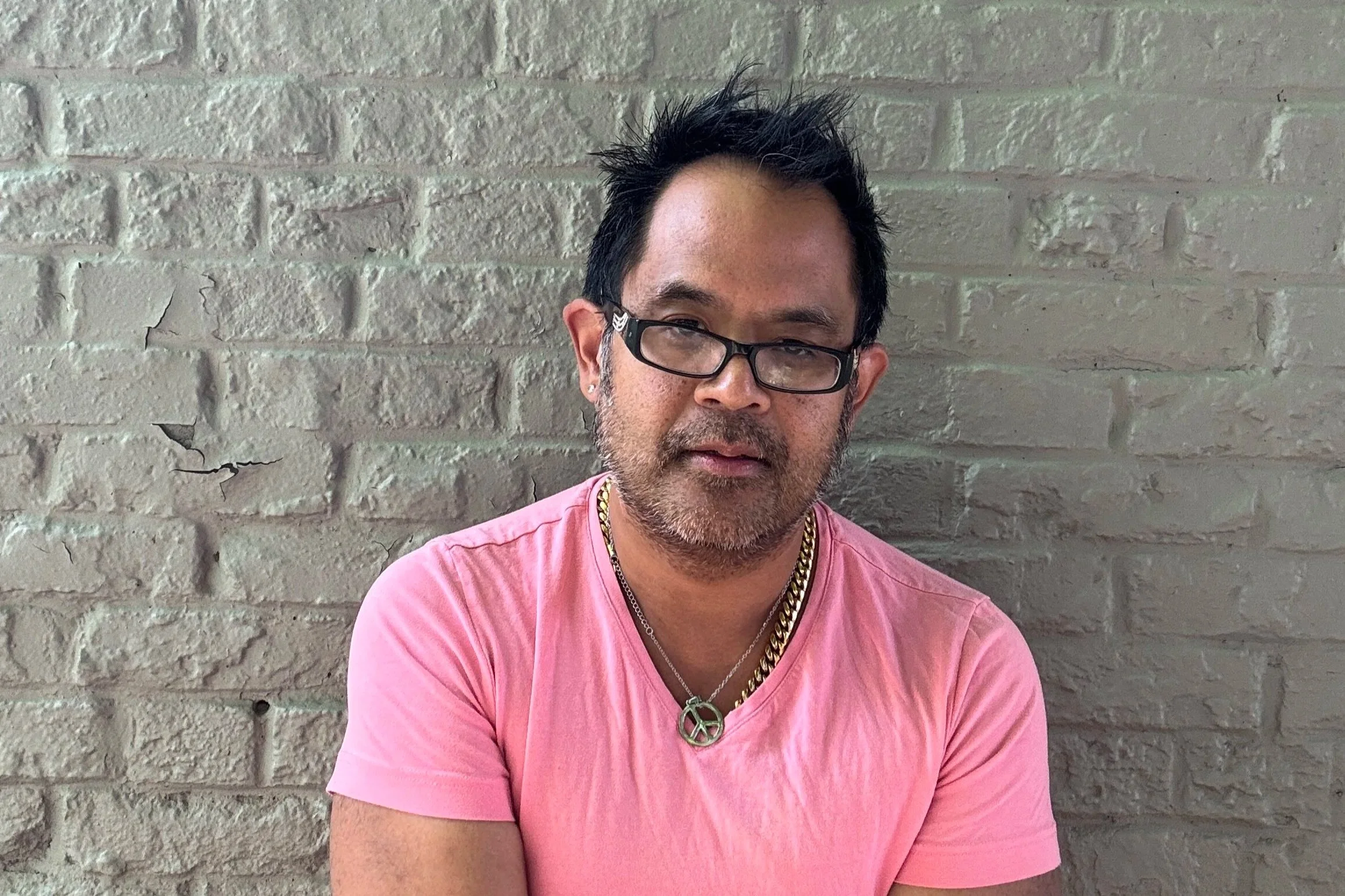 C.E. Gatchalian wearing glasses, a pink T-shirt, and layered necklaces posing in front of a gray brick wall