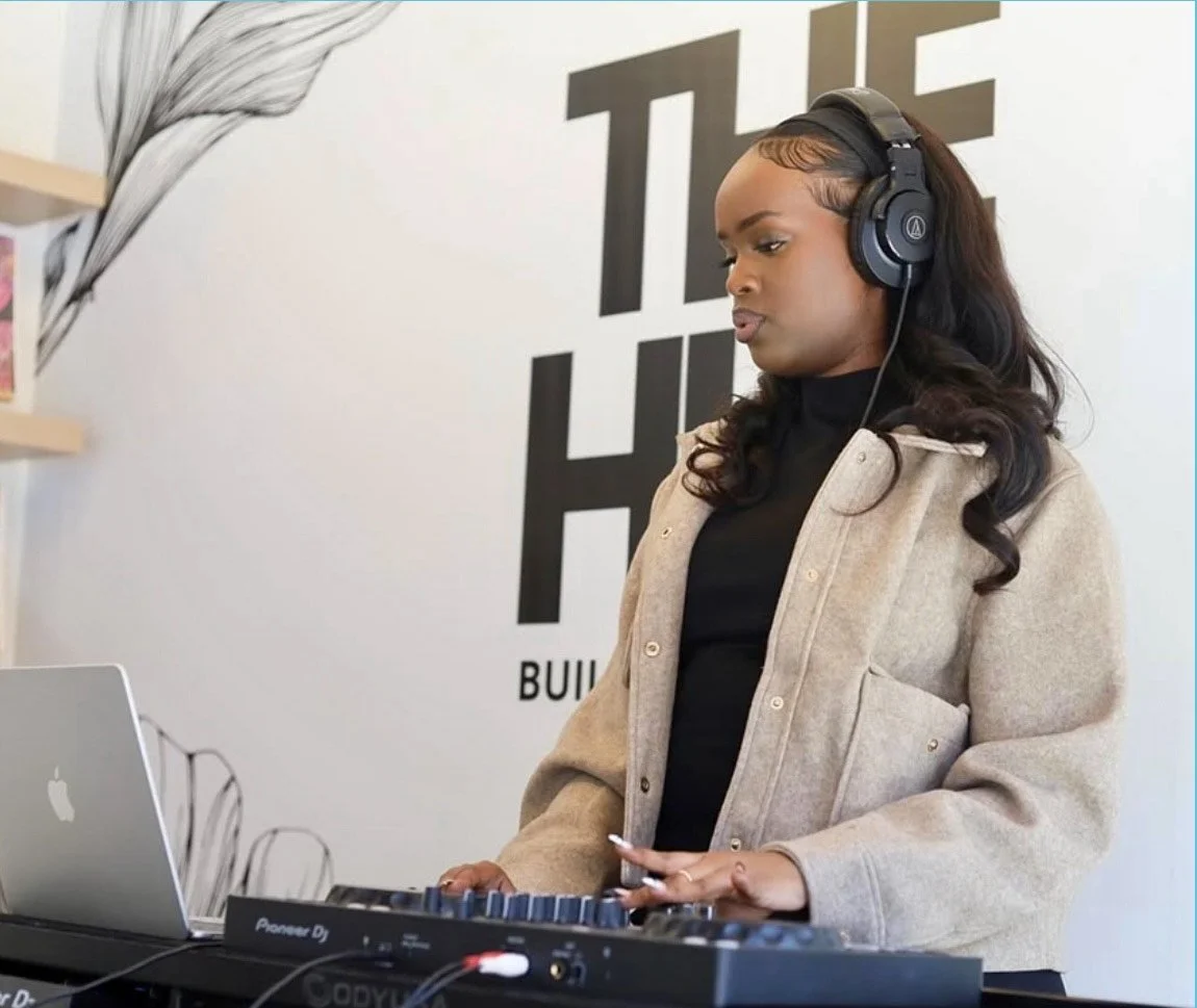 A woman with long, wavy hair listening to headphones and operating a DJ controller near a silver MacBook inside a modern building.