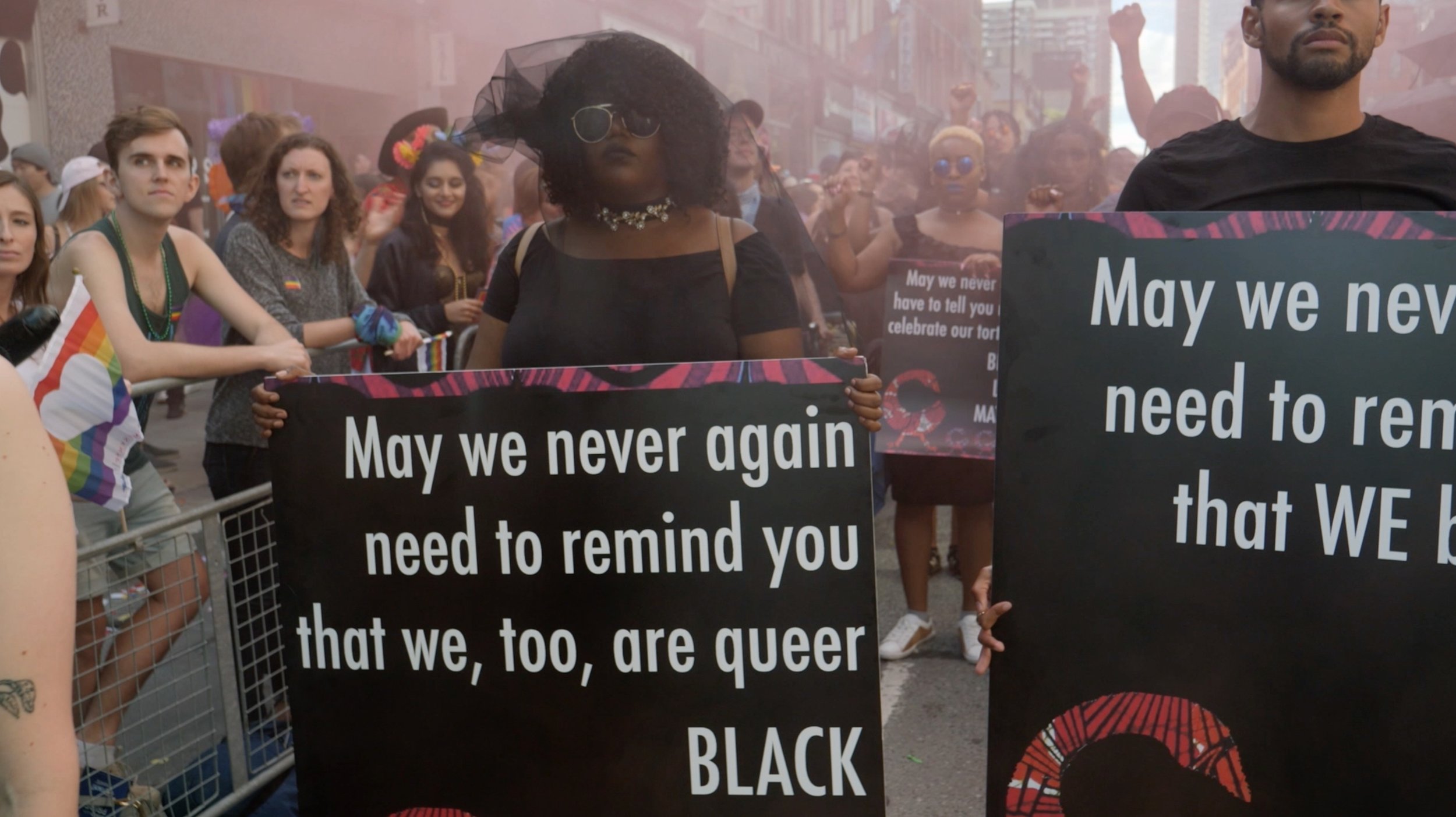 People participating in a pride parade holding signs with messages supporting Black queer individuals, surrounded by a crowd on a city street with fog or smoke in the background.