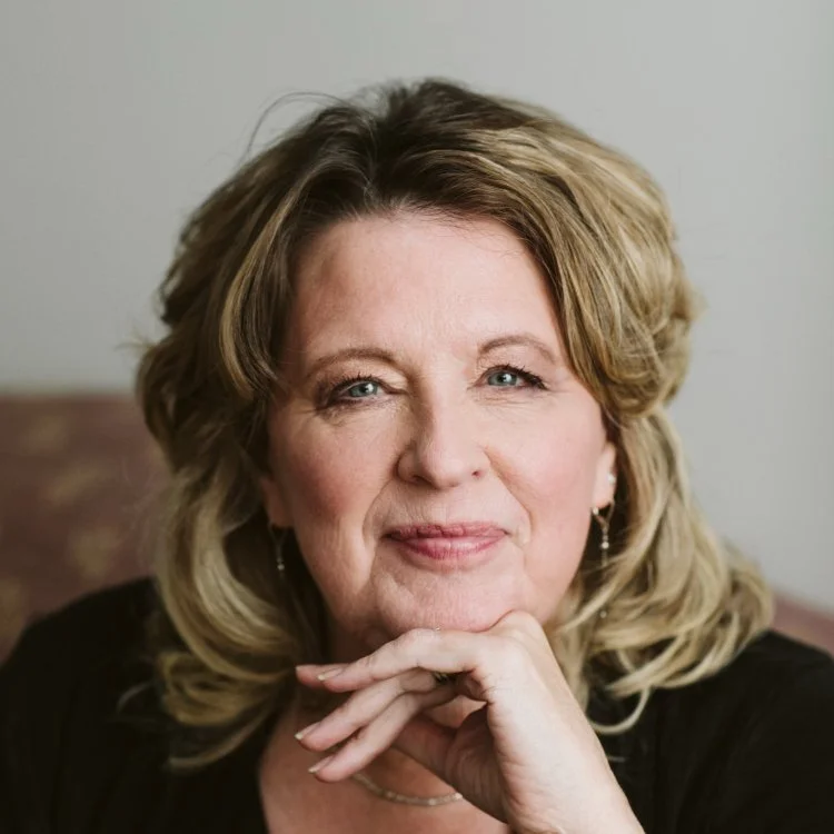 Portrait of a middle-aged woman with blonde hair, blue eyes, wearing earrings, and a black top, resting her chin on her hand, smiling softly. 2026 CCCU International Forum Speaker: Ruth Haley Barton.