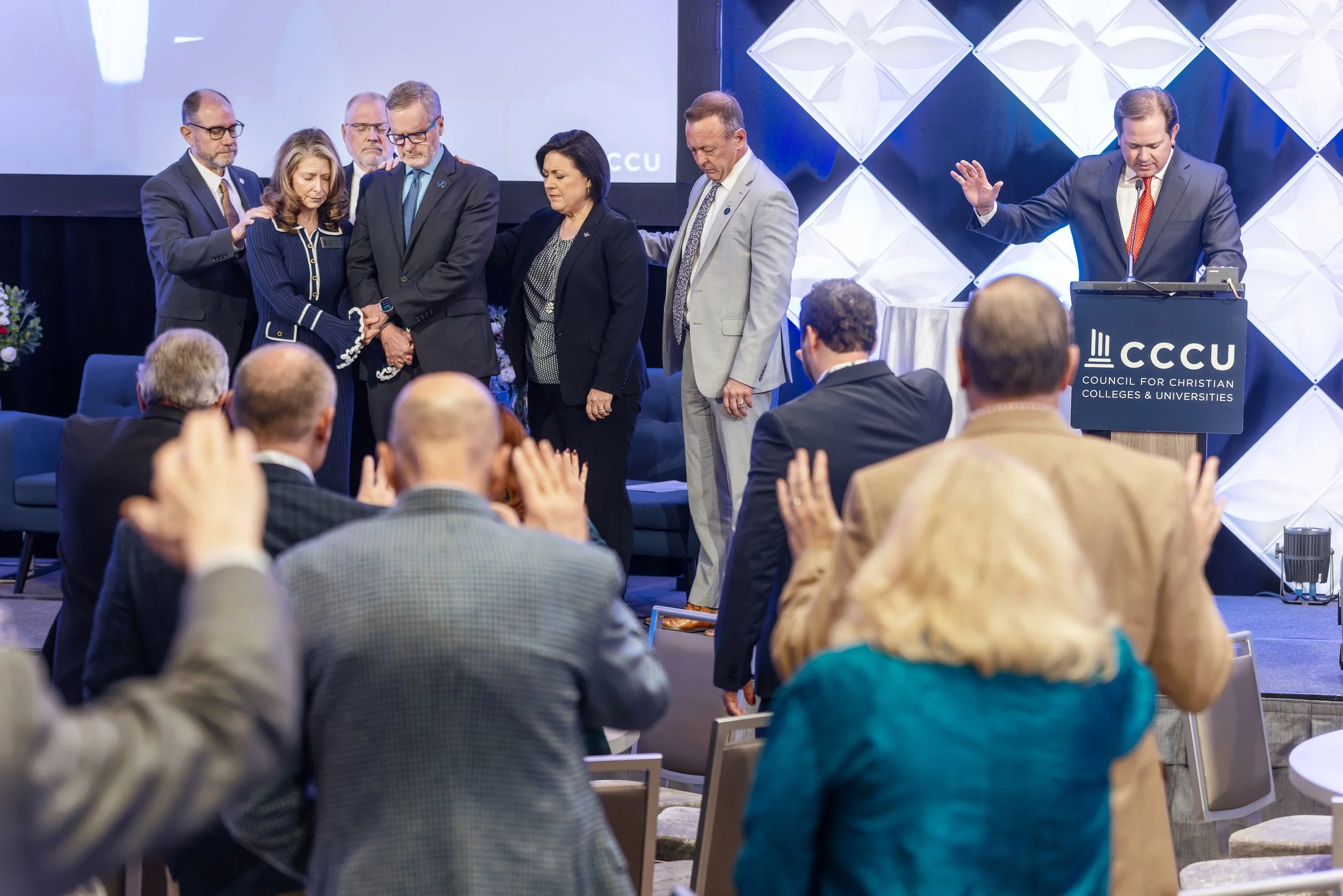 People standing on stage at a conference, some with heads bowed and hands clasped, with others in the audience applauding to celebrate 50 years of the CCCU from 1976 to 2026.